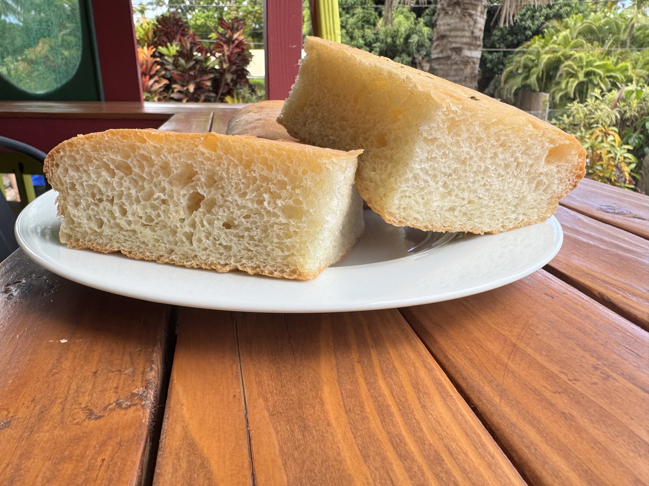 Close-up of sliced bread on a white plate, placed on a wooden table outdoors with greenery and trees in the background.