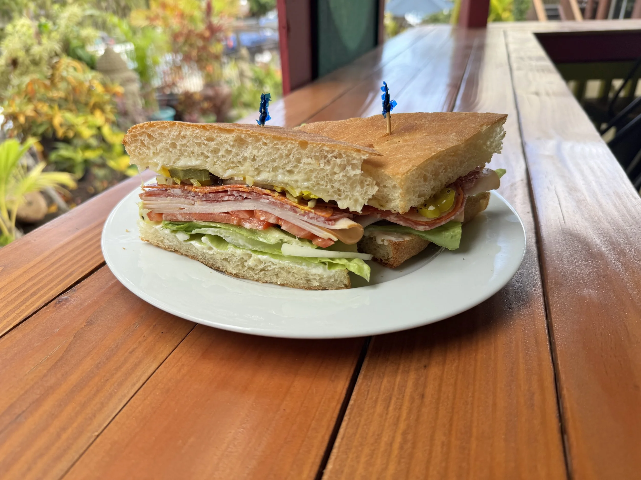 A sandwich on a white plate with layers of lettuce, tomato, sliced meats, and pickles, topped with two small blue flags, sitting on a wooden table outdoors.