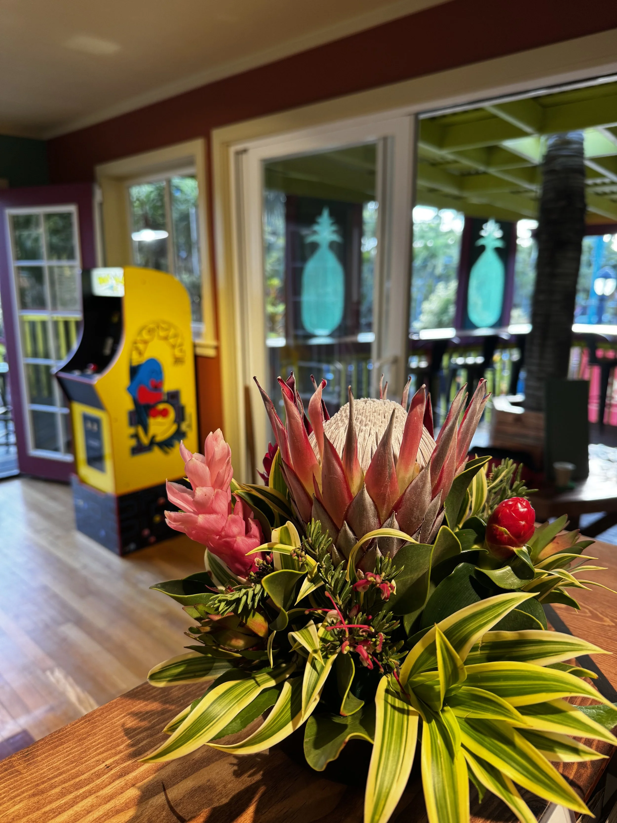 A potted plant with large green and yellow striped leaves, pink and red tropical flowers, and a central tall pineapple-shaped flower spike, placed on a wooden table in a cozy room.