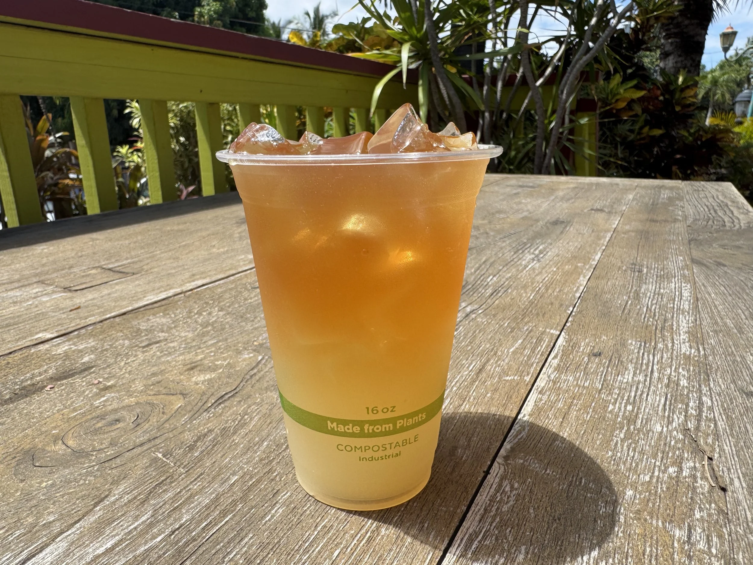 A plastic cup filled with an iced beverage on a wooden table outdoors, with a green railing and trees in the background.