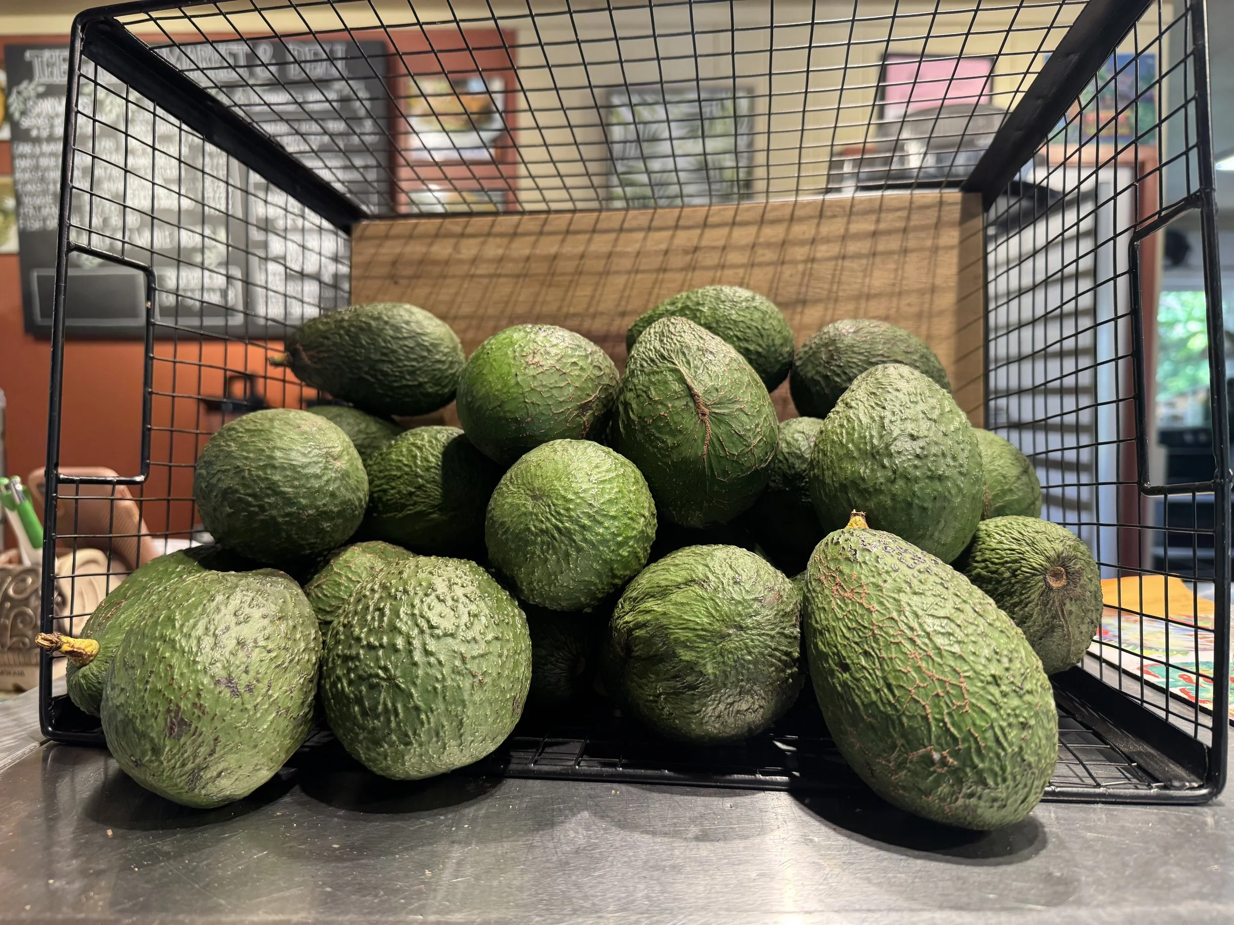 A shopping cart filled with green avocados inside a grocery store. The background shows a chalkboard menu and store decorations.