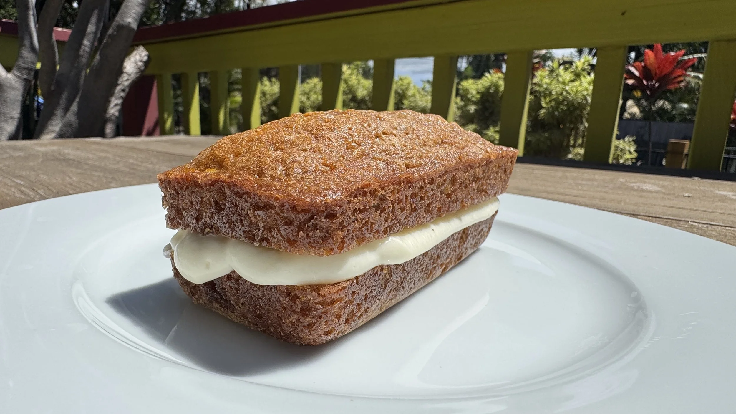 Close-up of a banana muffin with a slice of banana and cream cheese in the middle, on a white plate outdoors with a green painted wooden fence in the background.