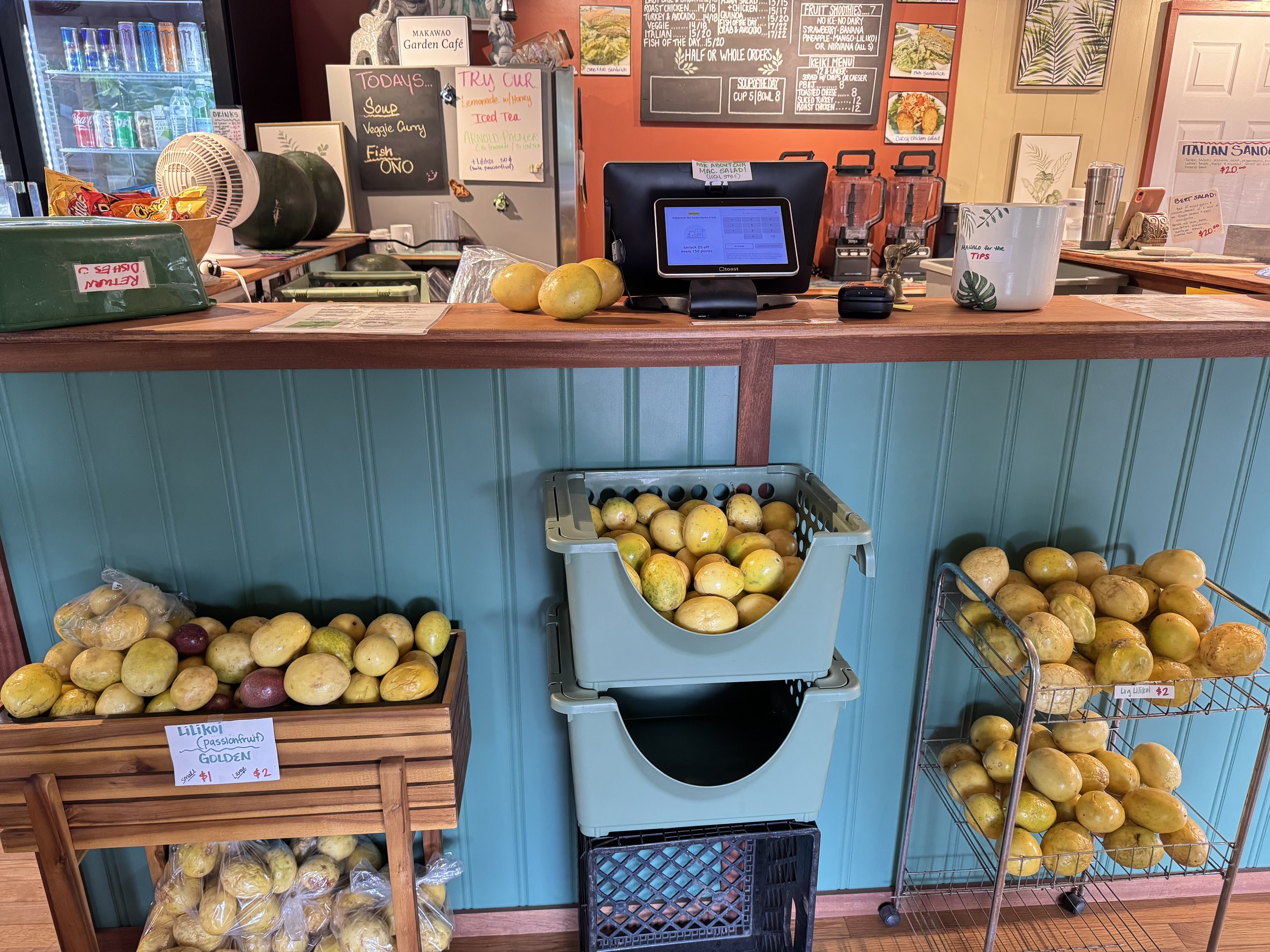 A wooden workspace with various fruits, a cash register, a glass of water, and a menu board. The fruits include apples and watermelons, placed in crates and a basket. There are signs with prices and product names, including passionfruit labeled 'Lili