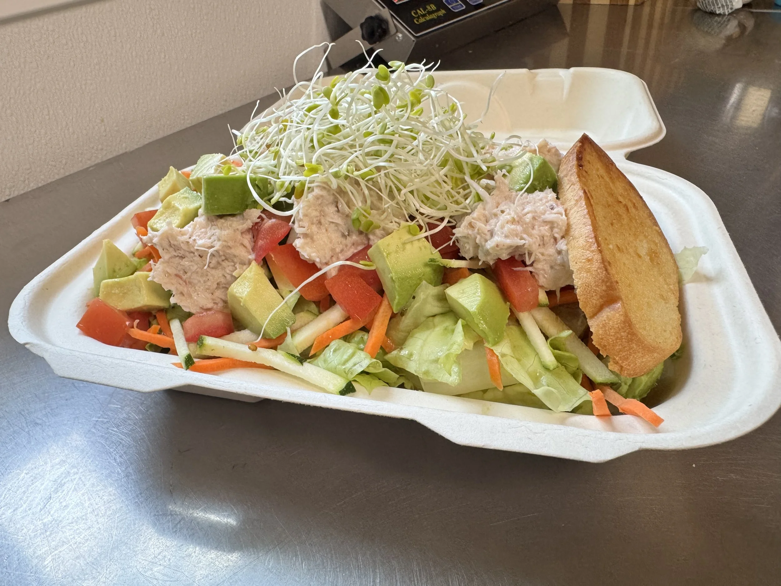 A takeout container filled with a salad that includes shredded lettuce, diced tomatoes, avocado chunks, shredded carrots, and a topping of microgreens, with a toasted bread slice on the side.
