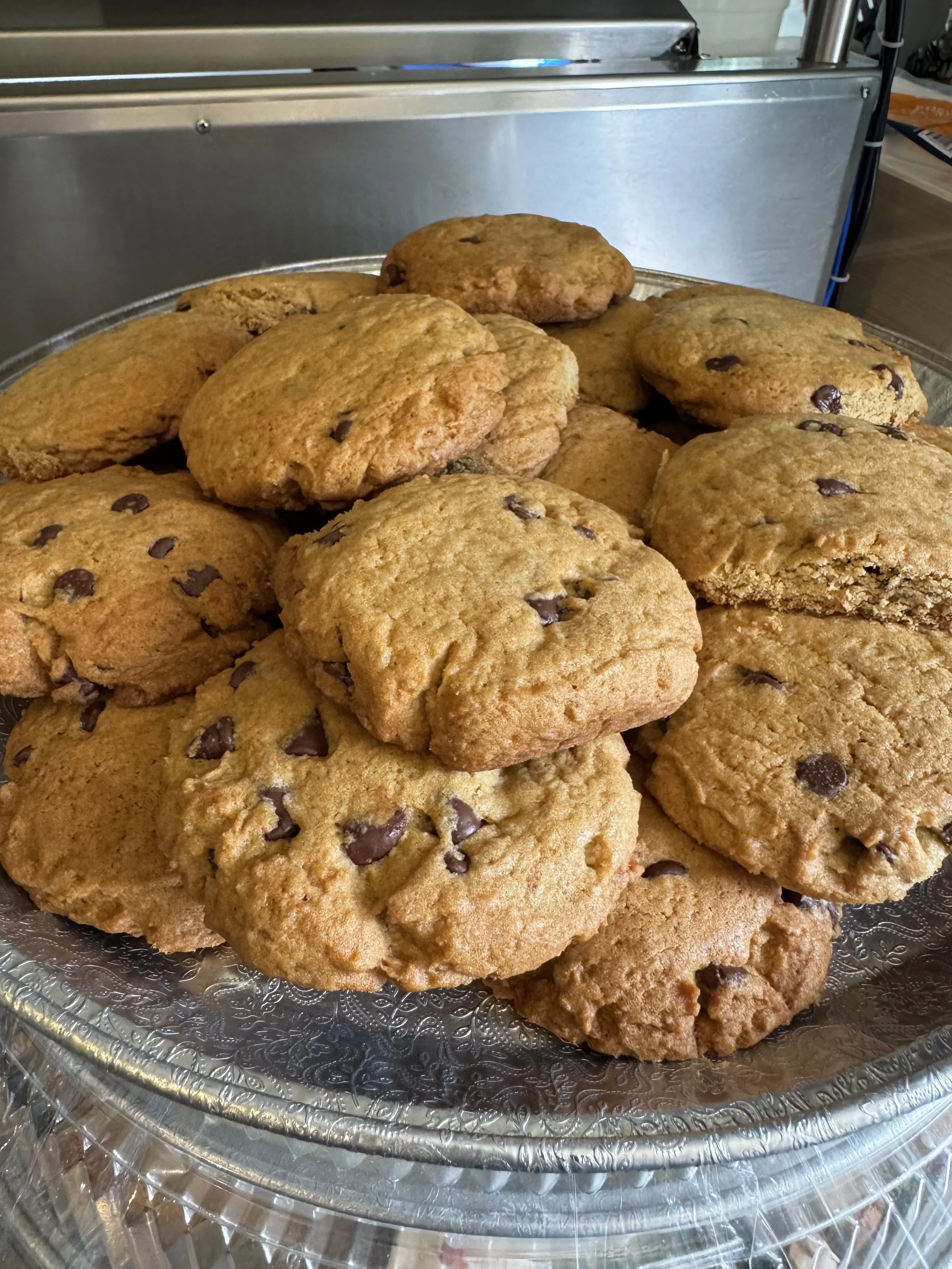 A silver tray filled with large chocolate chip cookies on a clear plastic sheet with a decorative rim. The background includes a metal structure and some partial view of a kitchen or food-preparation area.