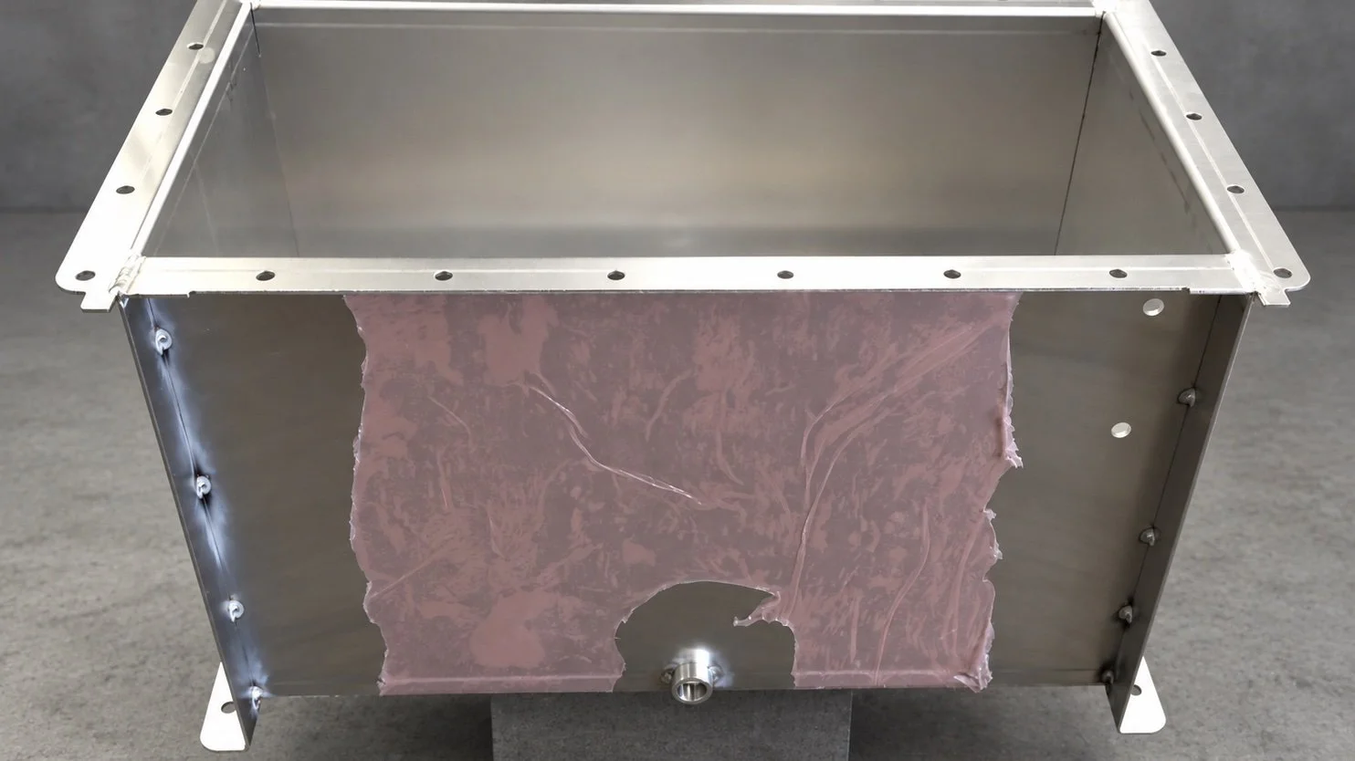 A metallic rectangular container with a pinkish substance inside, visible on one side, with a small metal knob at the bottom center of the front panel.