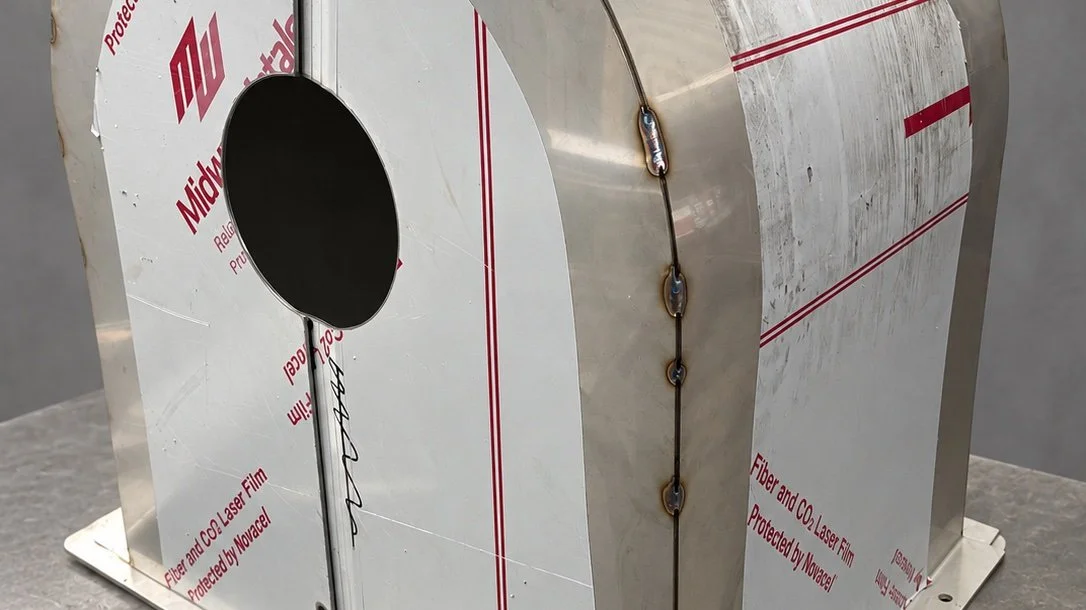 A metal cylindrical vessel with welds and protective film partial removal, sitting on a gray surface.