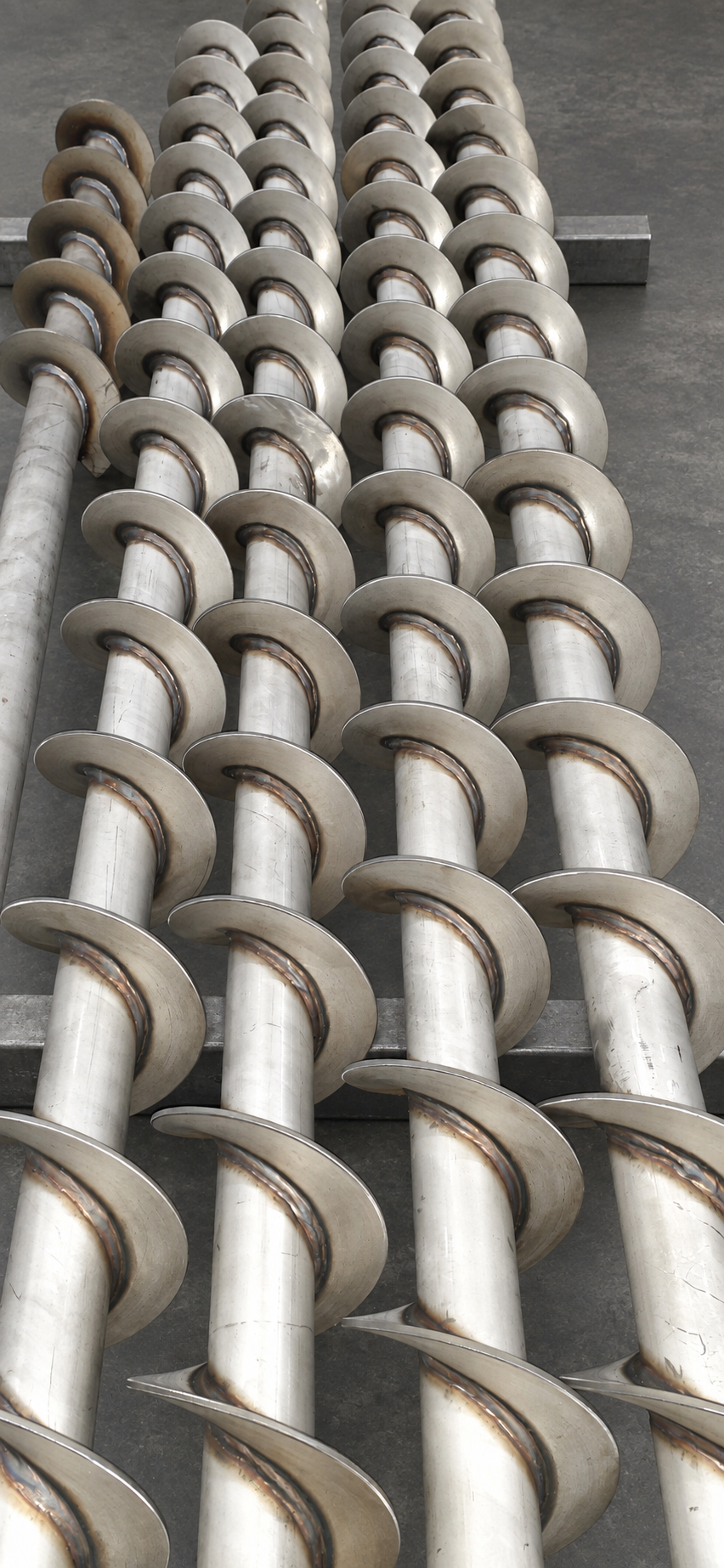Industrial metal screw conveyor shafts arranged in parallel on a concrete floor.