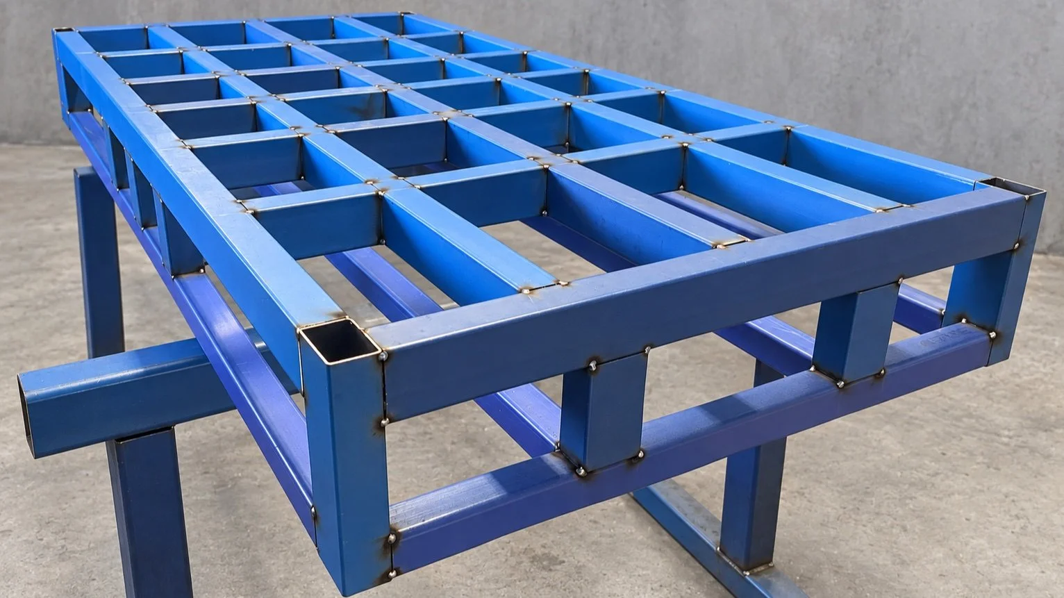 A blue metal workbench or frame with a grid-like top structure, made of rectangular tubes welded together, placed on a concrete floor against a gray wall.