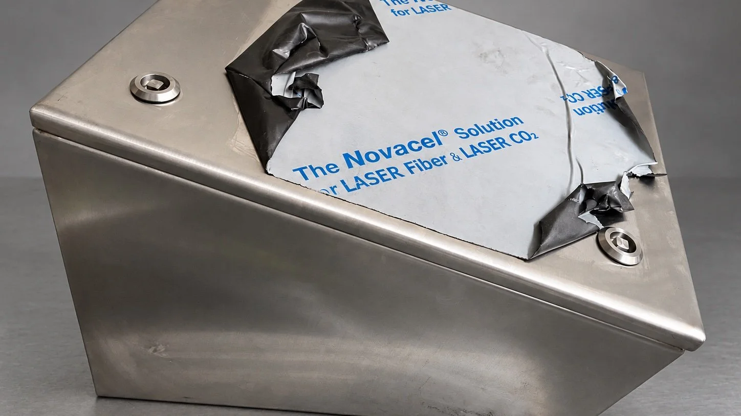 A stainless steel container with a torn label that reads "The Novacel Solution for LASER Fiber & LASER CO2." The lid material is peeled back, revealing the inside of the container.
