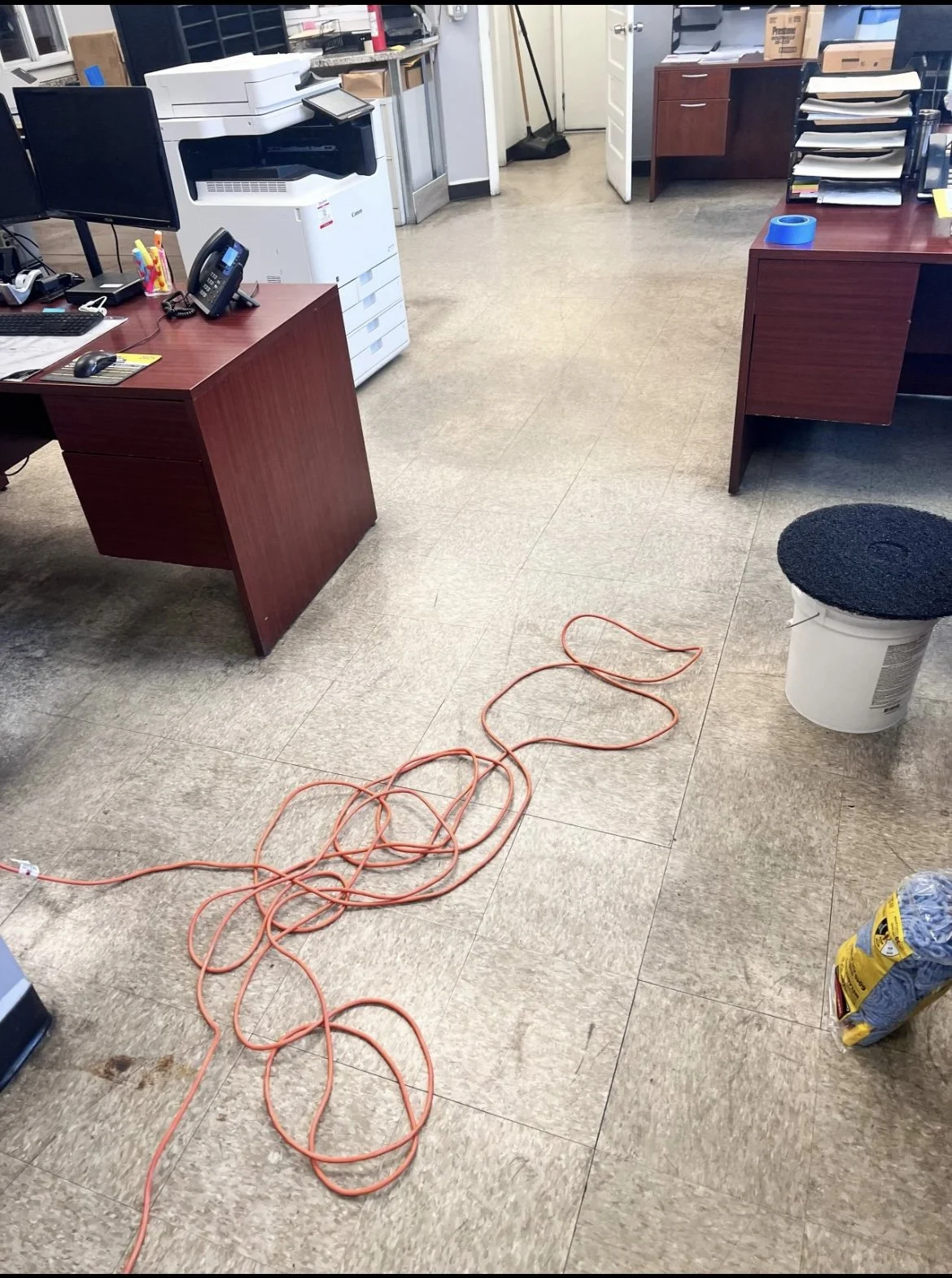 An office with wooden desks, computers, and office supplies. There is an orange extension cord on the floor, a white trash bin with a black lid, and cleaning tools in the background.