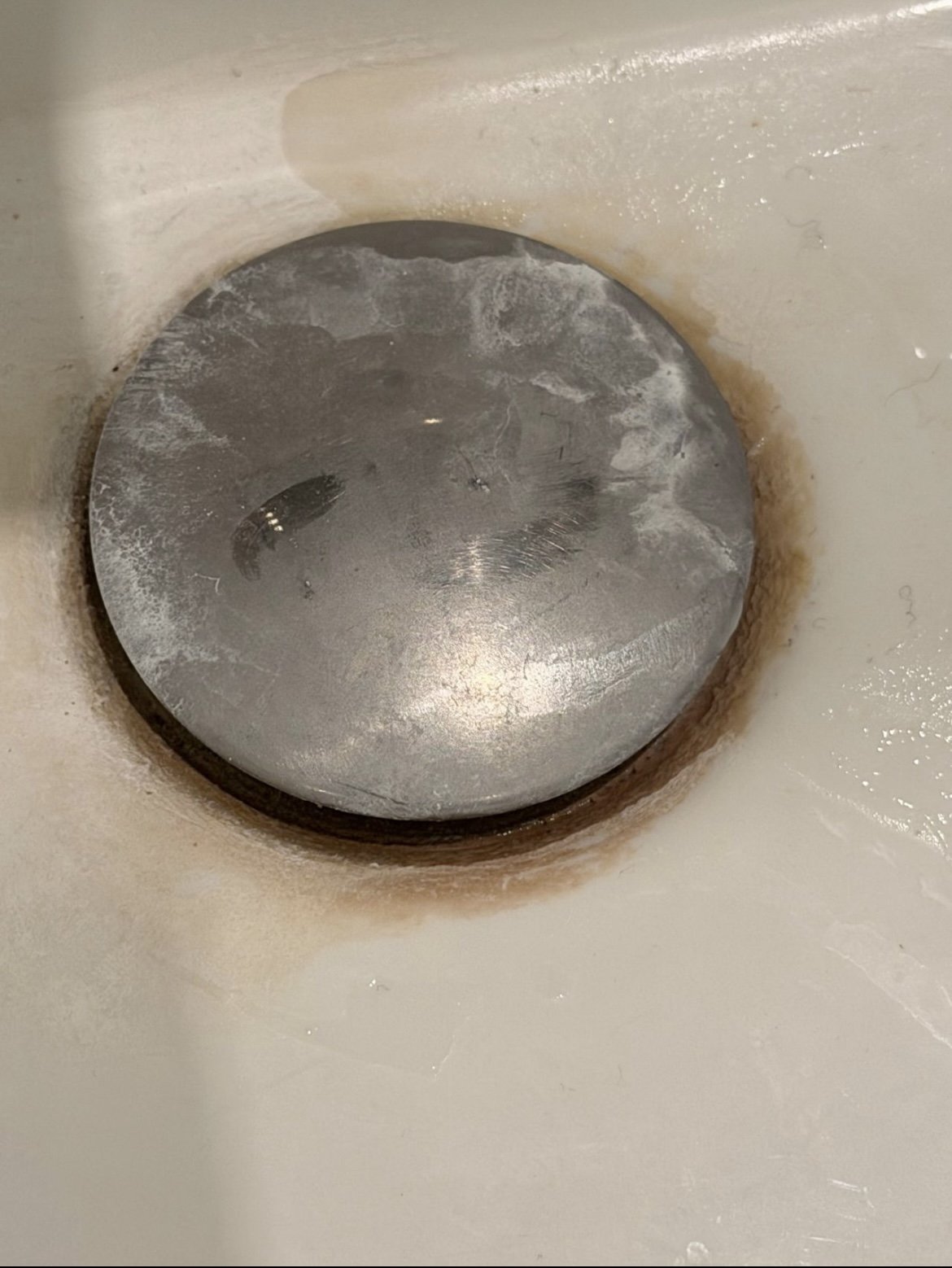 Close-up image of a metal drain plug inside a bathroom sink with signs of rust and residue around the drain opening.