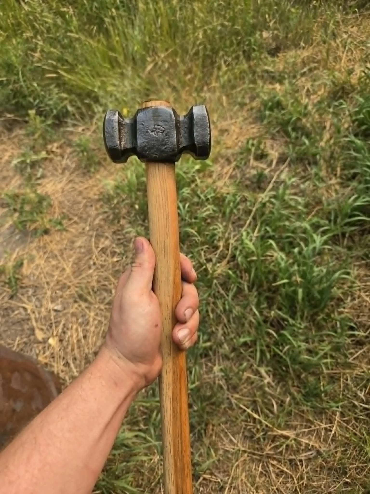 A person's hand holding a hammer with a wooden handle over grass and dirt ground.