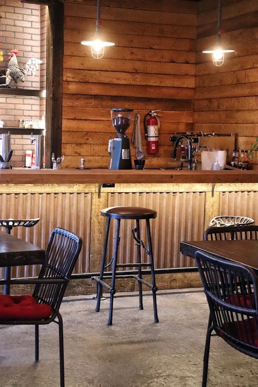 Interior of a cozy cafe or bar with wooden walls, black chairs with red cushions, a wooden bar counter, and three hanging pendant lights.