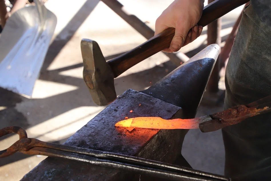 Blacksmith forging a piece of metal on an anvil, with tools in hand, in a workshop setting.