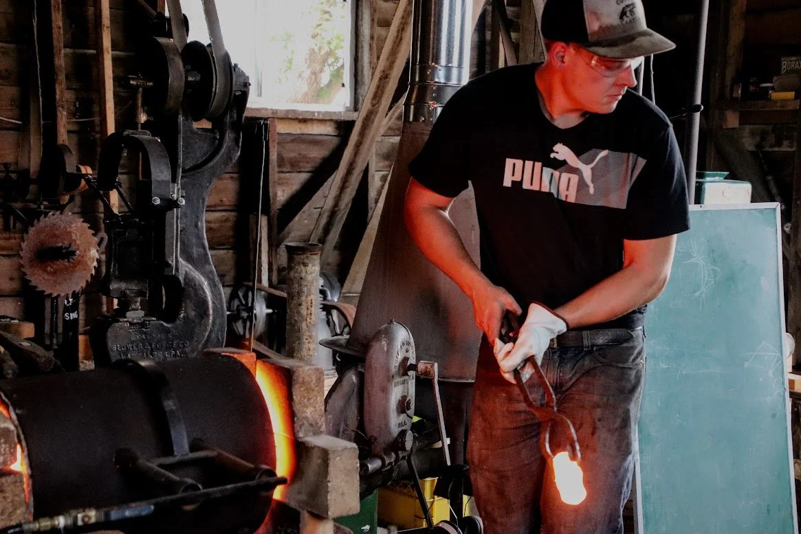 A man in a black Puma t-shirt and a cap working with glowing heated metal in a blacksmith workshop.