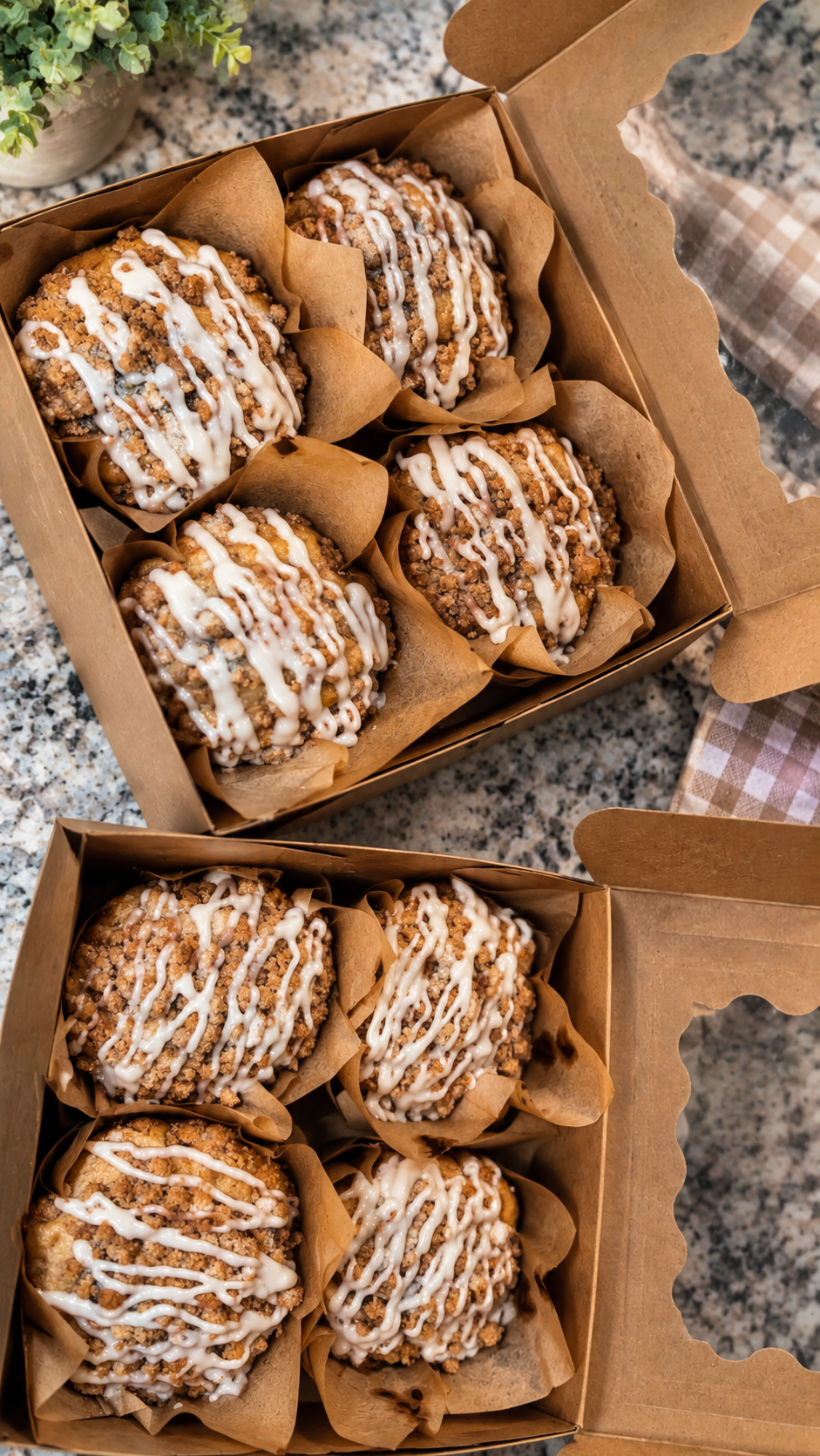 Two open boxes of crumbly muffins drizzled with white icing, lined with parchment paper, resting on a granite countertop with a small plant and a checkered napkin nearby.