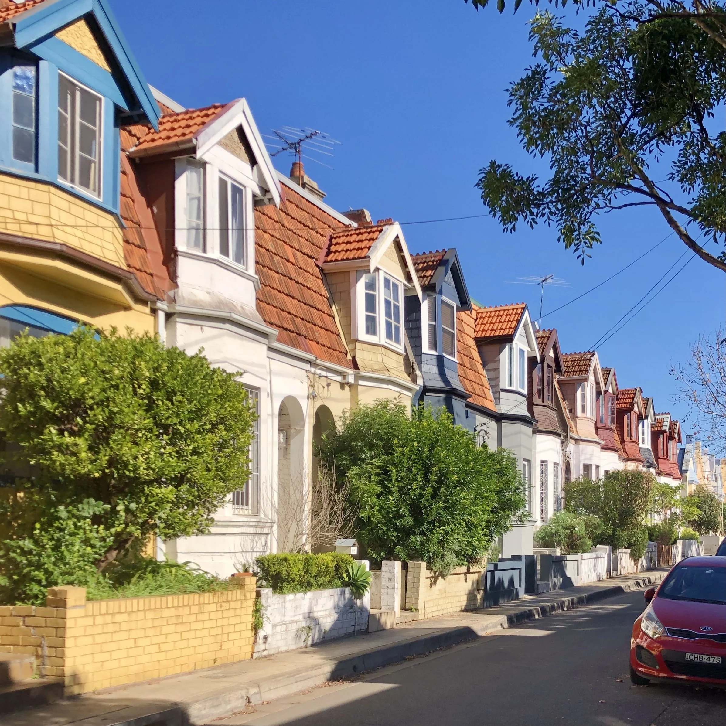 A row of colorful connected houses with bay windows, red-tiled roofs, and a brick wall in front, along a street with parked cars and trees under a clear blue sky.