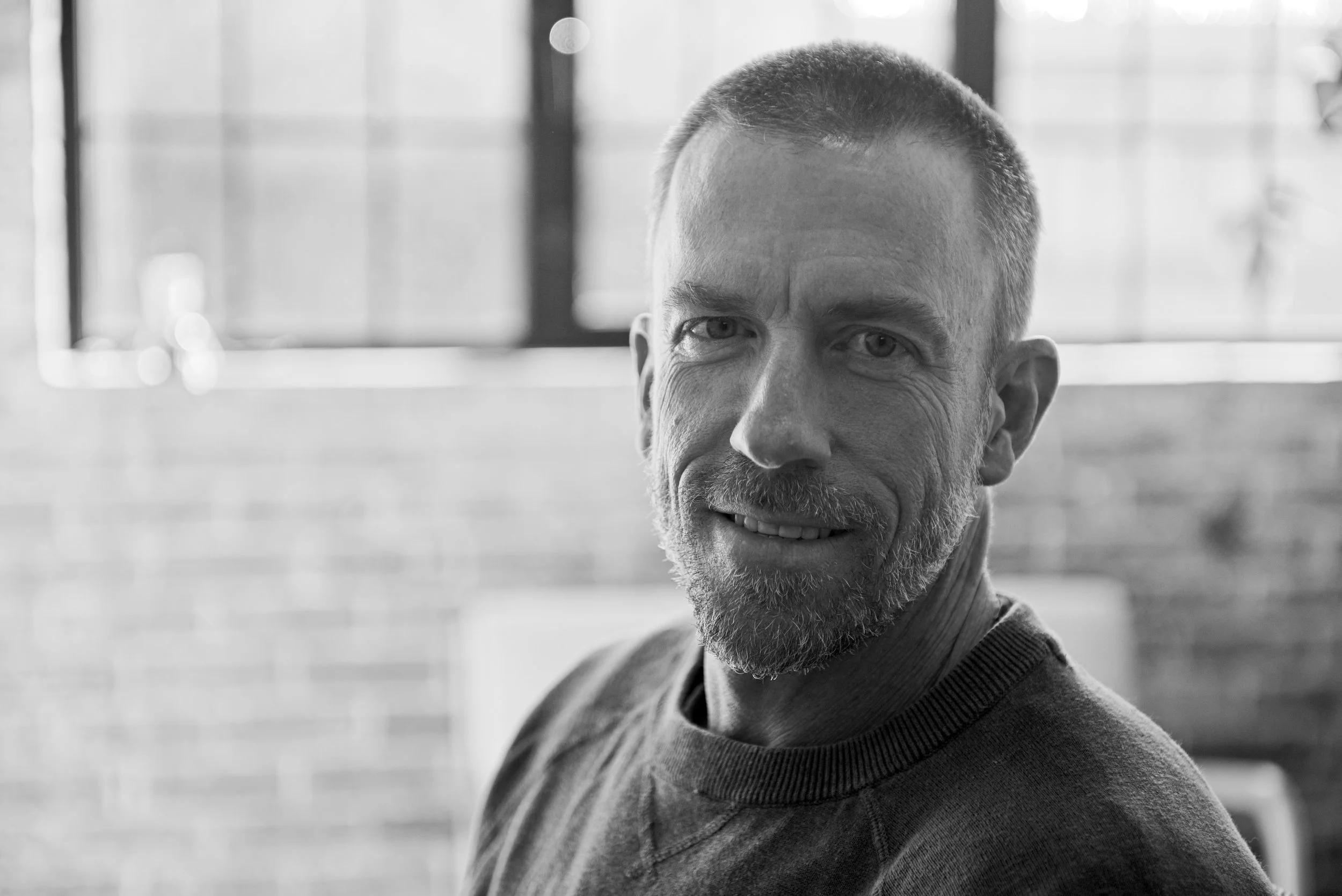 A black-and-white photo of a smiling middle-aged man with short hair and a beard, wearing a sweater, sitting indoors near large windows with blurred background.