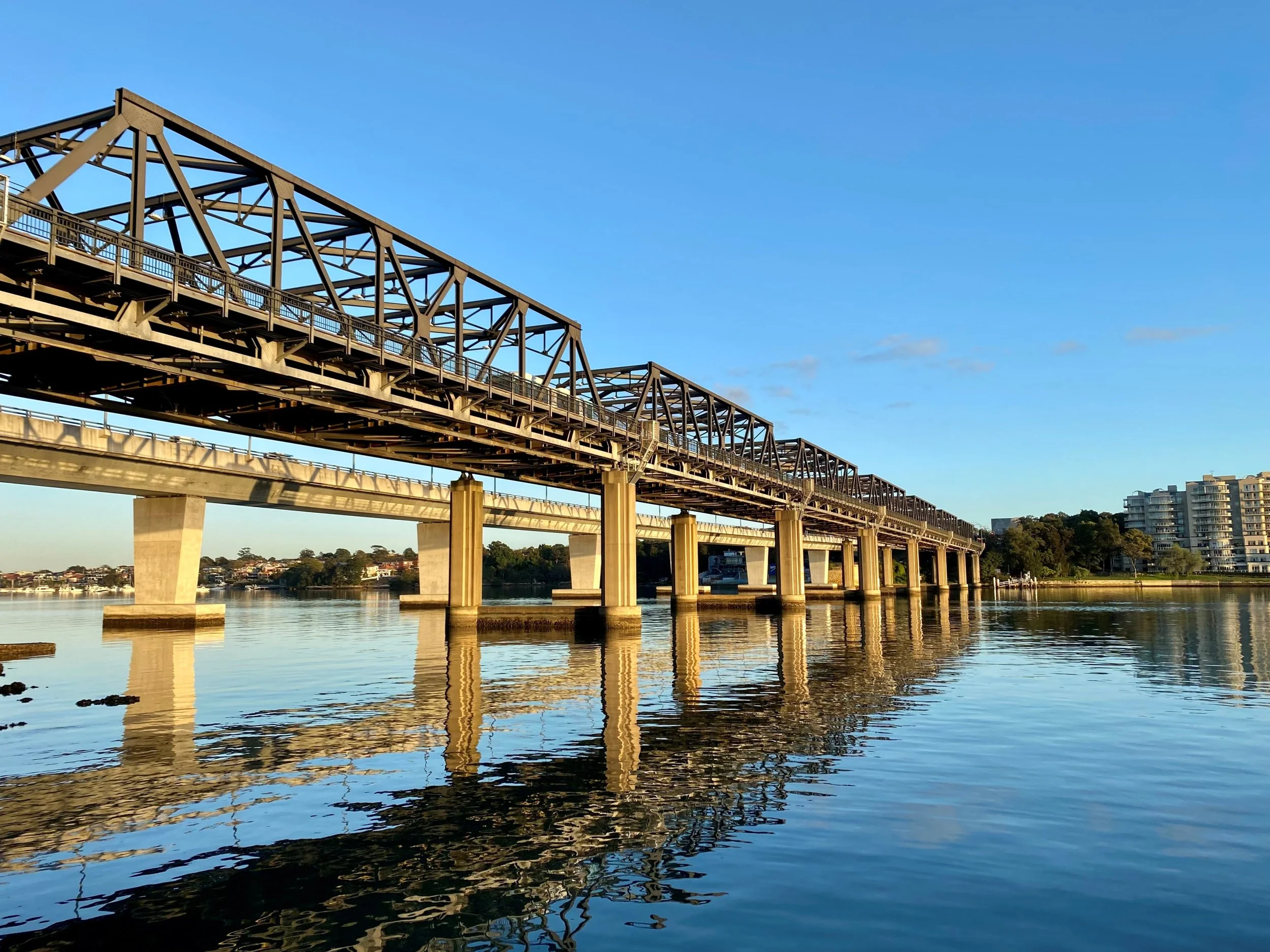 Steel bridge extending over calm water with buildings and trees in the background under a clear blue sky.