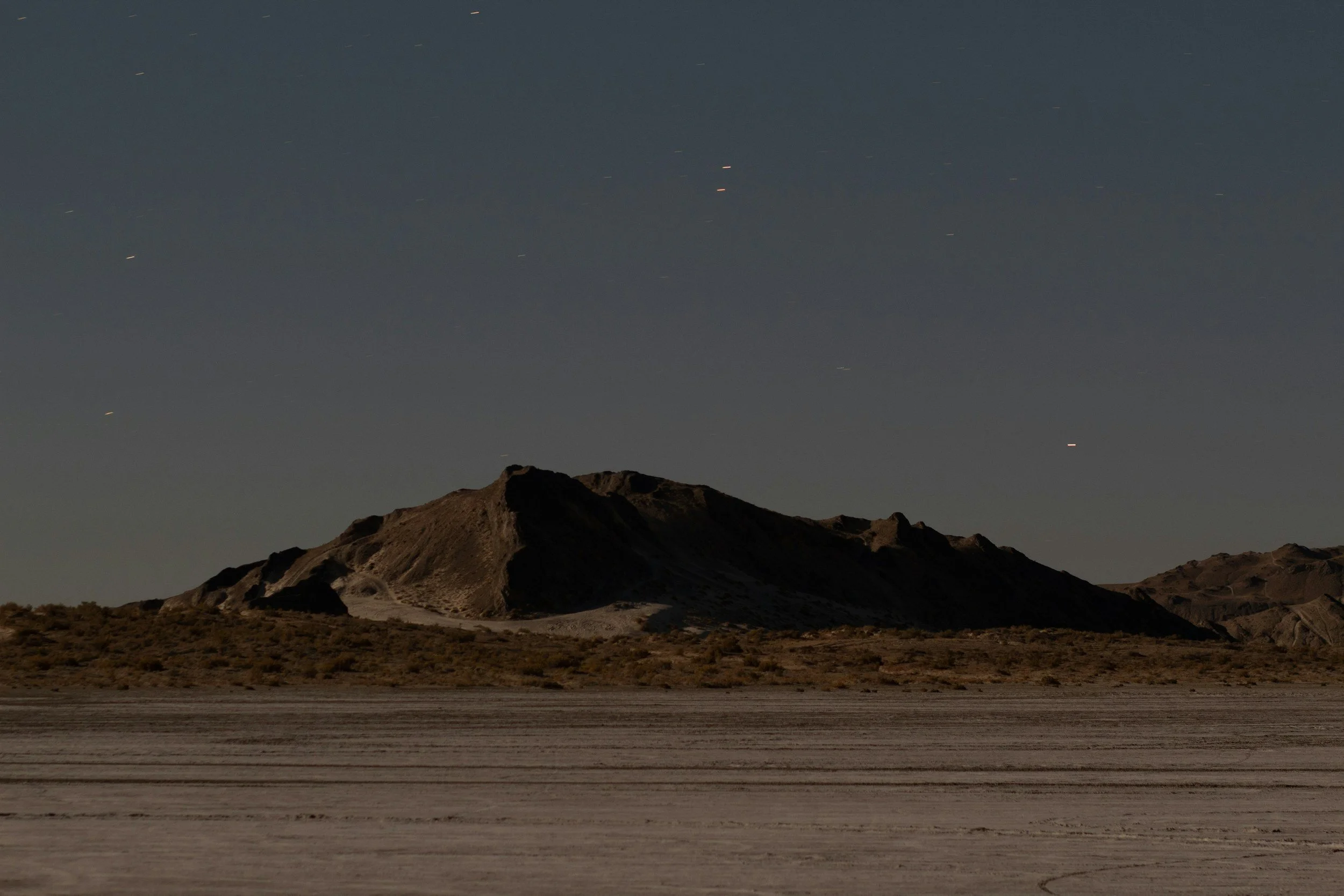Nighttime landscape with a rugged mountain in the background and a wide, flat desert foreground. The sky is clear with faint star trails.