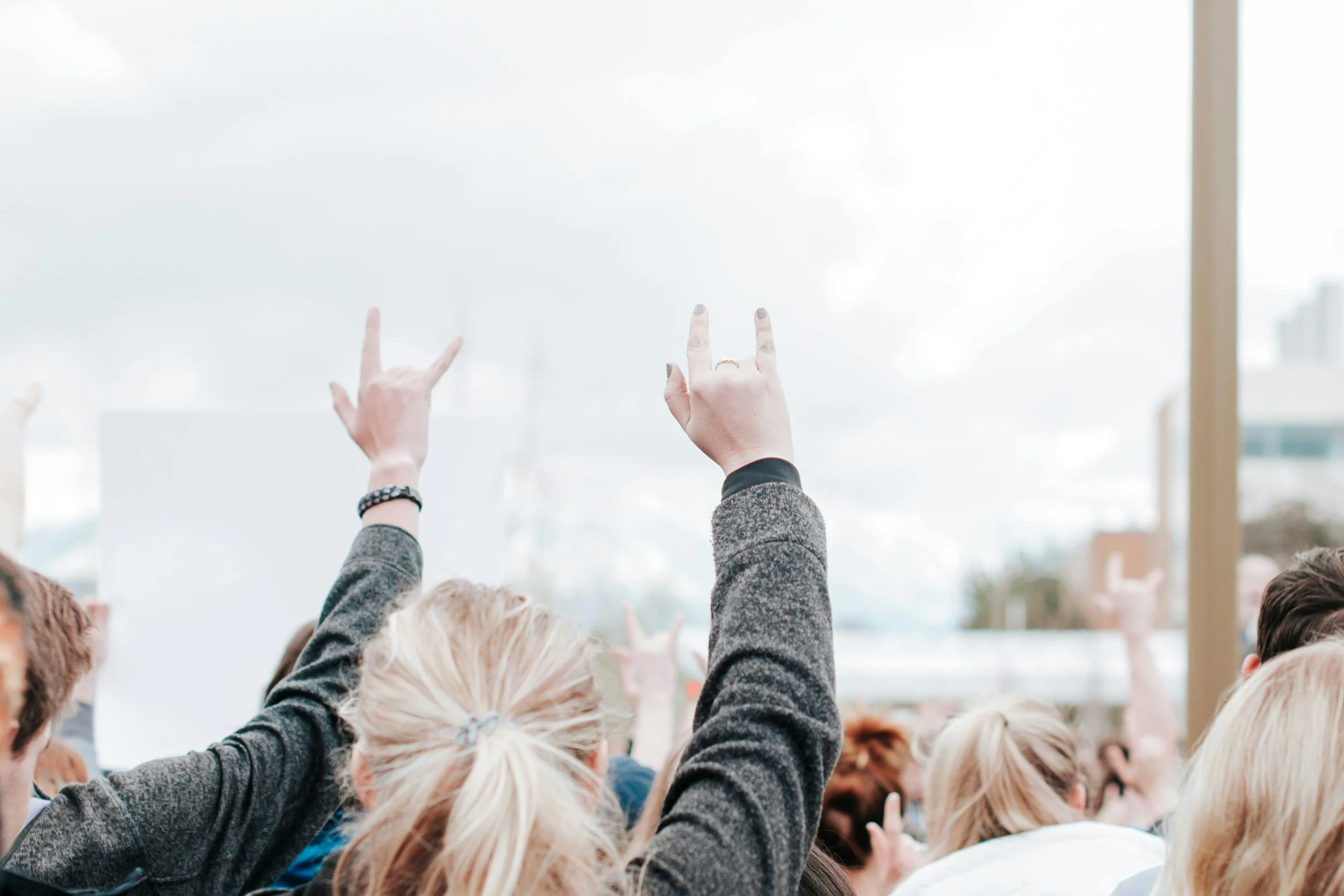 Crowd of people raising their hands at a gathering or event, with some making the rock and roll hand sign, indoors with a large window and city buildings visible in the background.