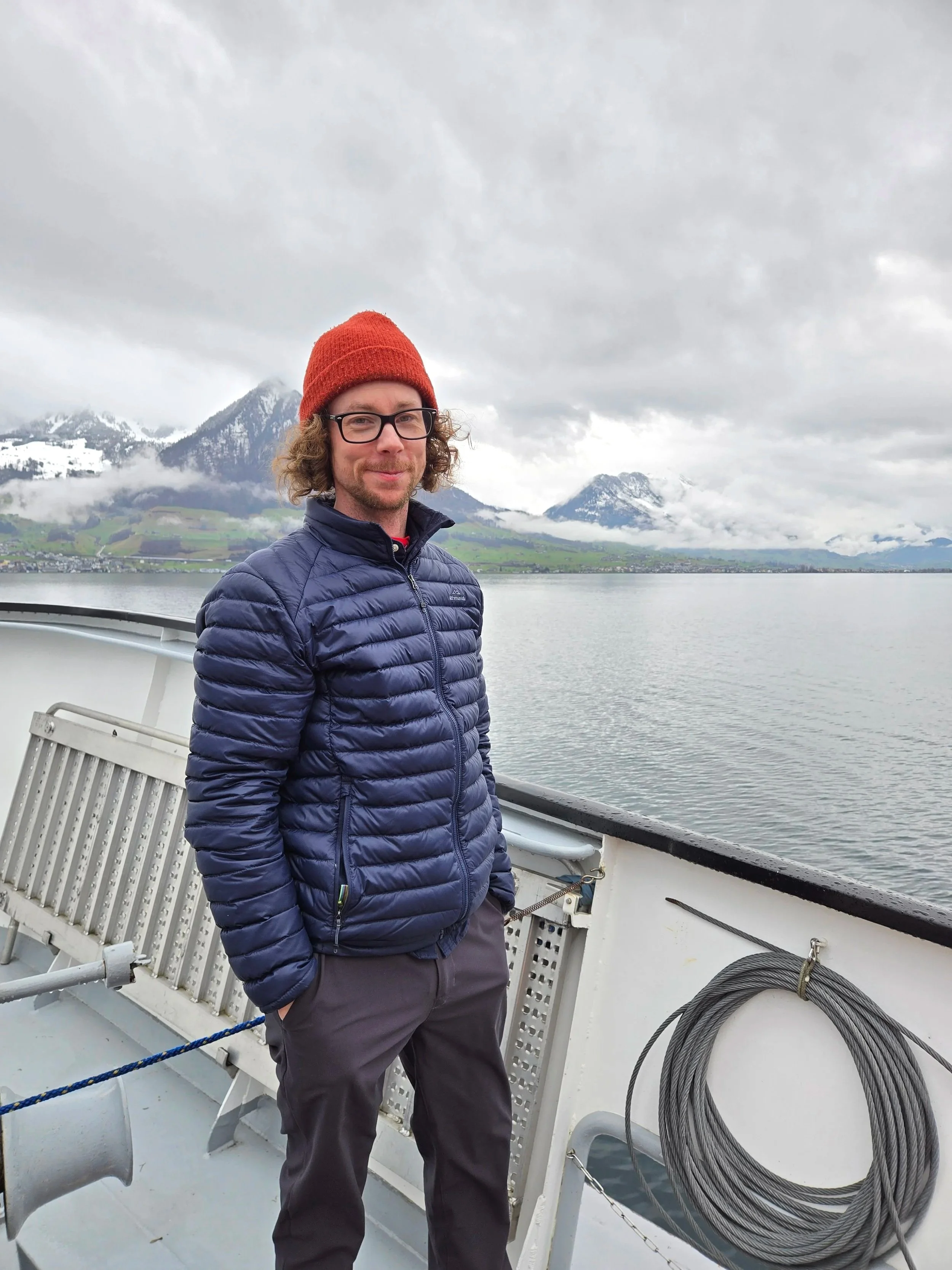 A man with glasses and a red knit cap standing on a boat with mountains and a cloudy sky in the background.
