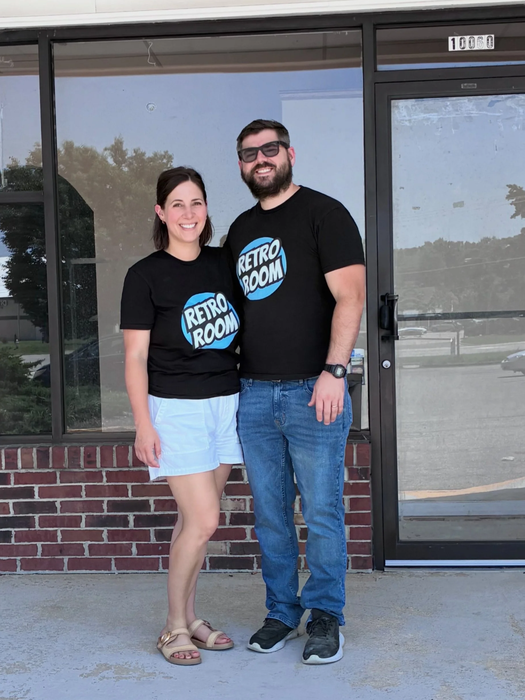 A man and a woman standing outside in front of a glass door, both wearing black t-shirts with a blue and white logo that says 'RETRO ROOM', smiling at the camera