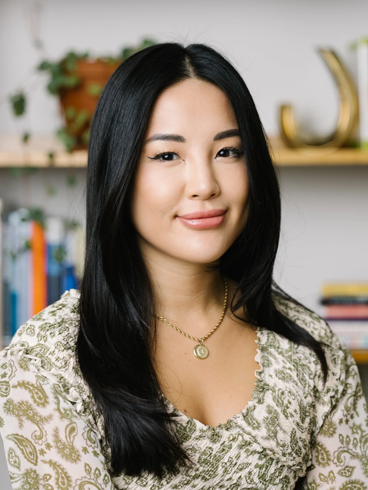 Portrait of a woman with long black hair in a patterned top, wearing a gold chain necklace with a pendant, standing in an indoor setting with books and plants in the background.