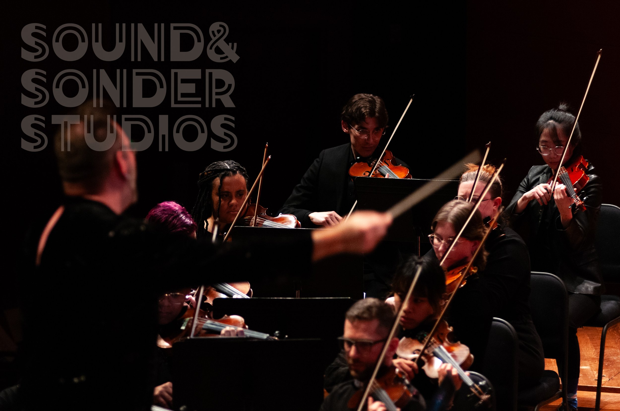 Orchestra performing on stage with musicians playing violins and other string instruments, with a dark background and warm lighting.