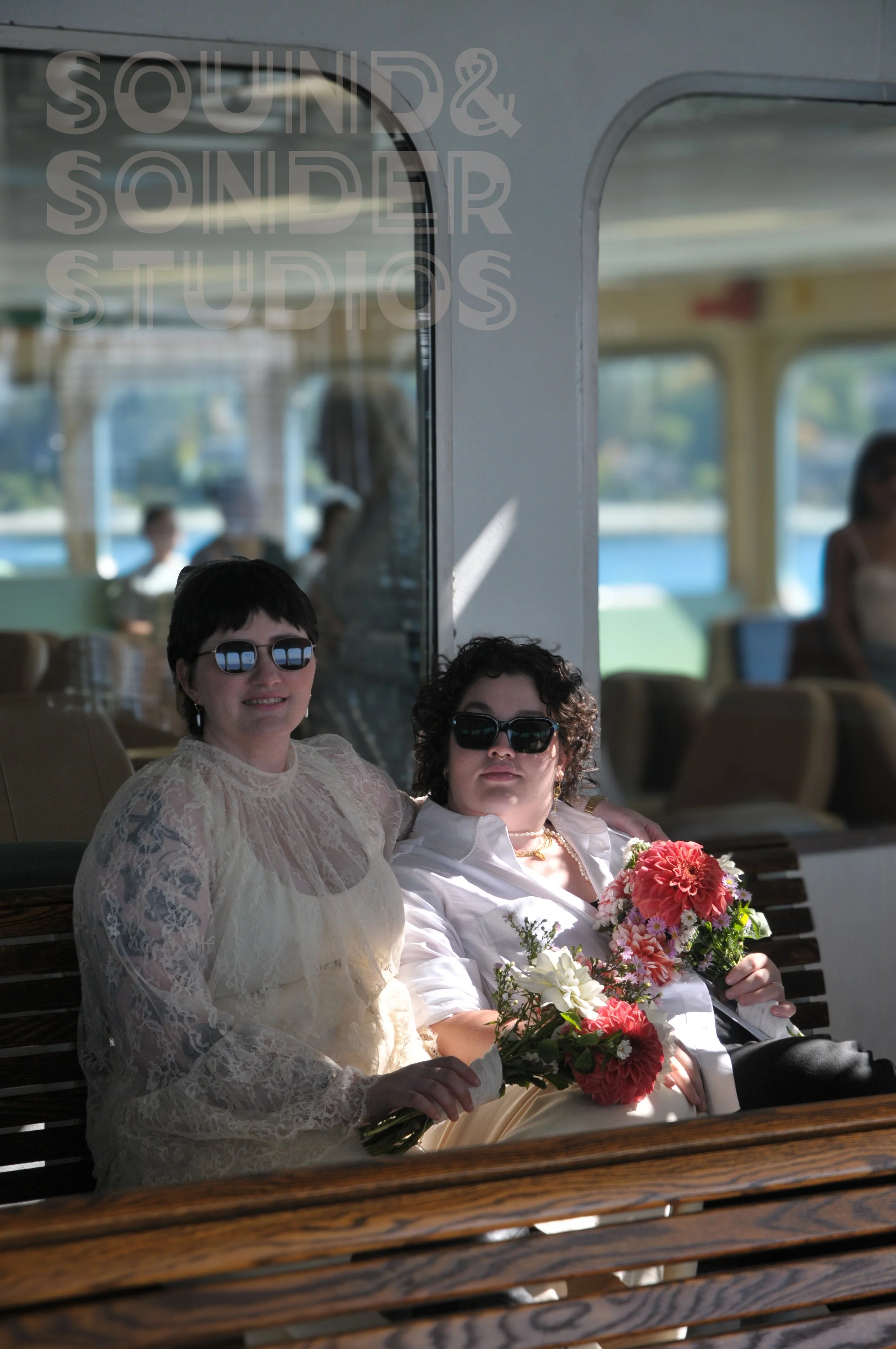Two women with dark sunglasses sitting on a wooden bench with a woman holding a bouquet of pink and white flowers. Behind them, the interior of a boat with windows showing a water view.