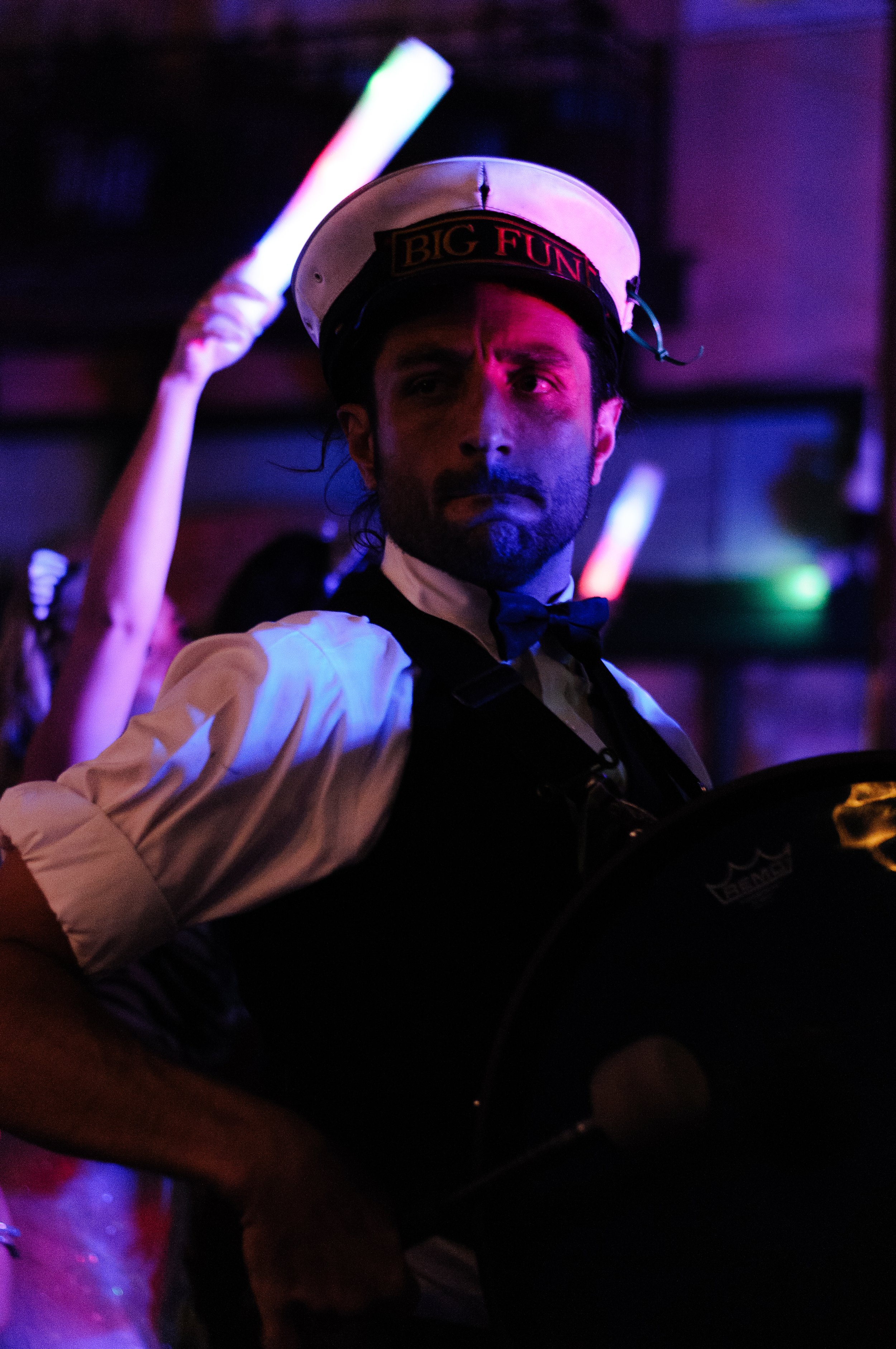 A man dressed as a funeral director or bartender, wearing a white shirt, black vest, and bowtie, is in a dark, colorful club or event space. He wears a white hat with the words "BIG FUN" on it, and is looking into the camera. He is holding a glow stick or neon light in his raised hand, with purple, green, and pink lighting effects in the background.