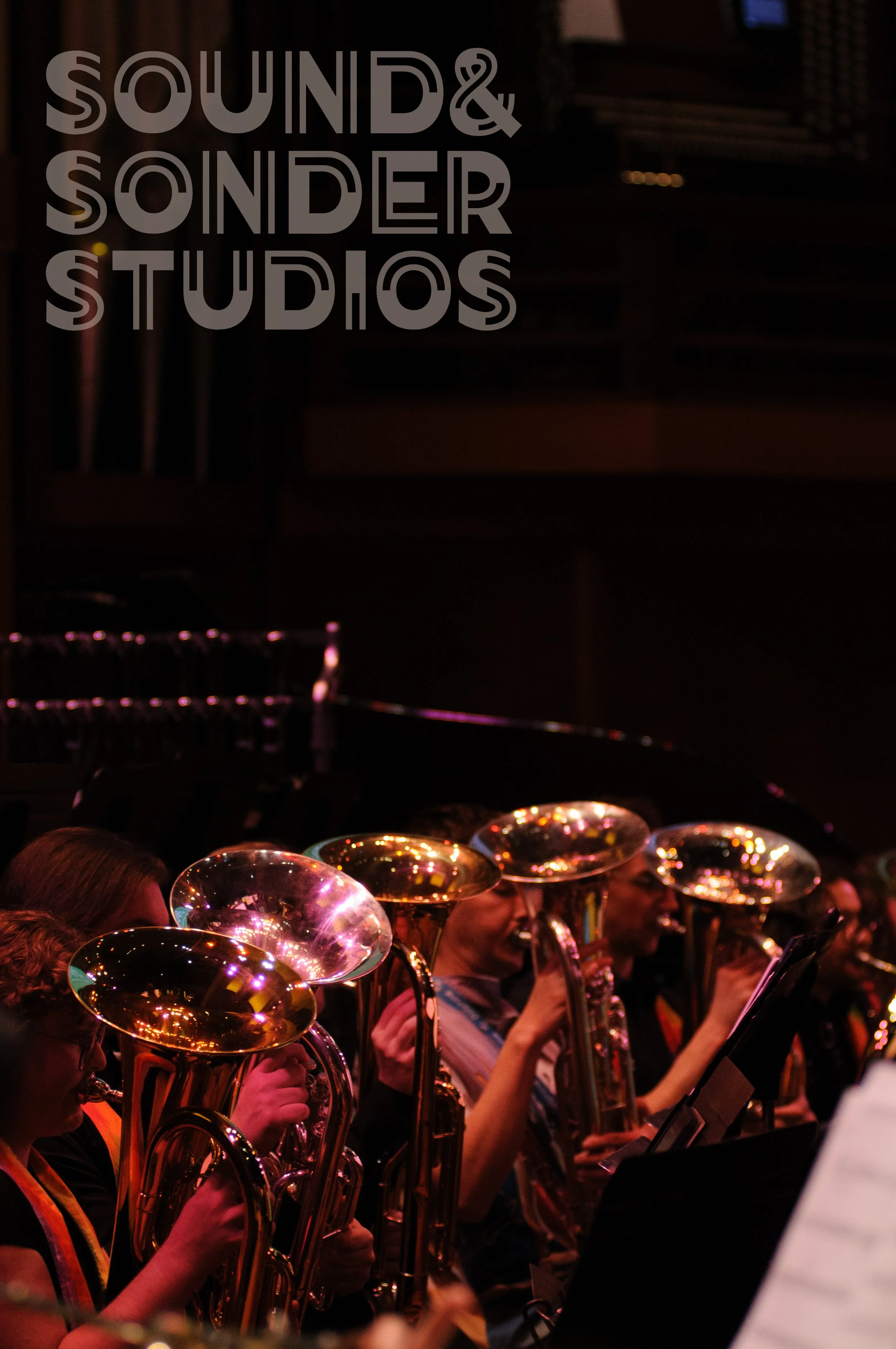 A group of musicians playing saxophones during a performance in a dimly lit venue. Some of the musicians are wearing orange vests, and they are reading sheet music. The background has a sign that reads 'Sound & Sounder Studios'.