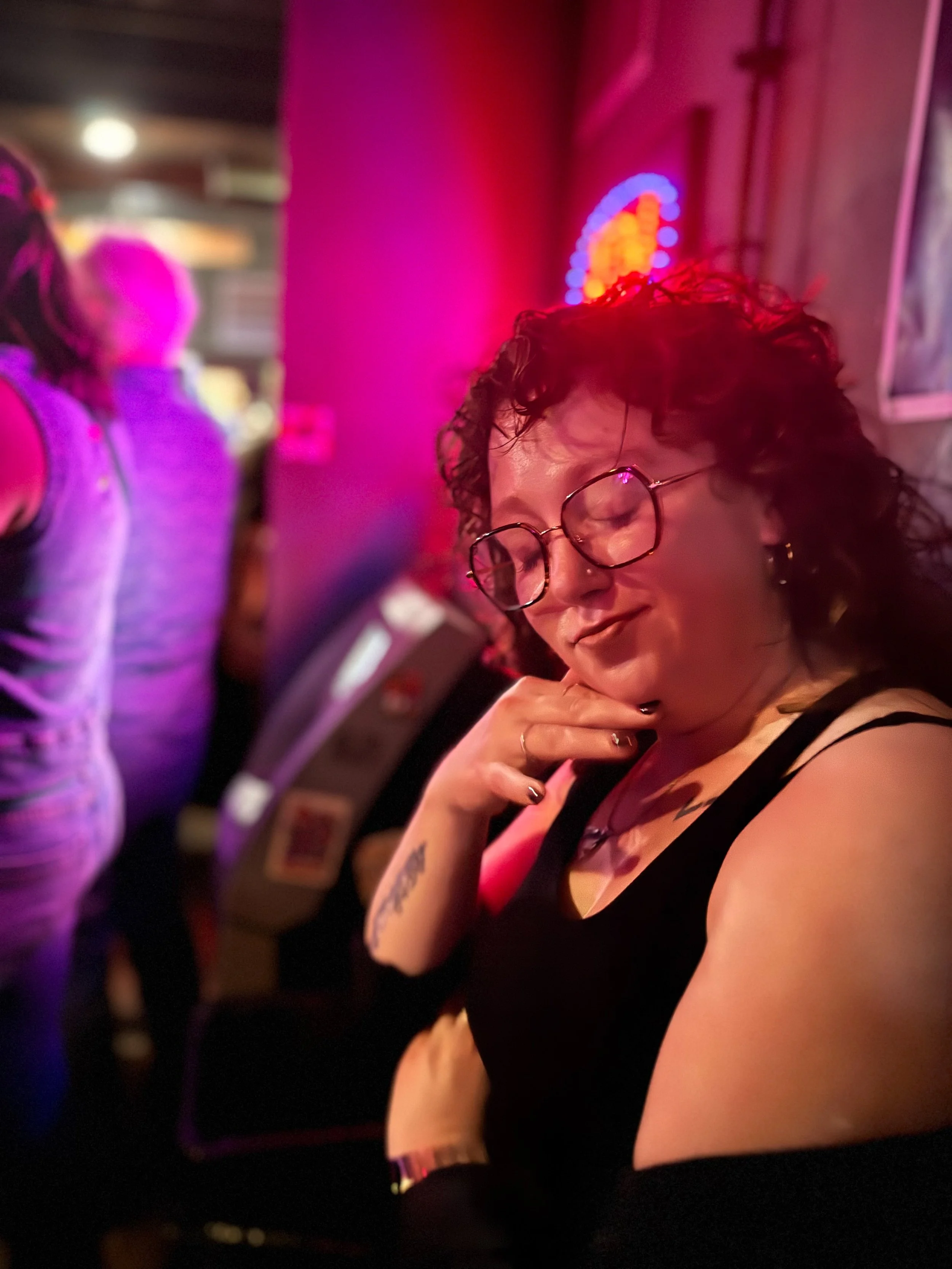 A woman with curly hair and round glasses is in a dimly lit room with pink and purple neon lighting, wearing a black sleeveless top. She is touching her chin with one hand and has tattoos on her arm.