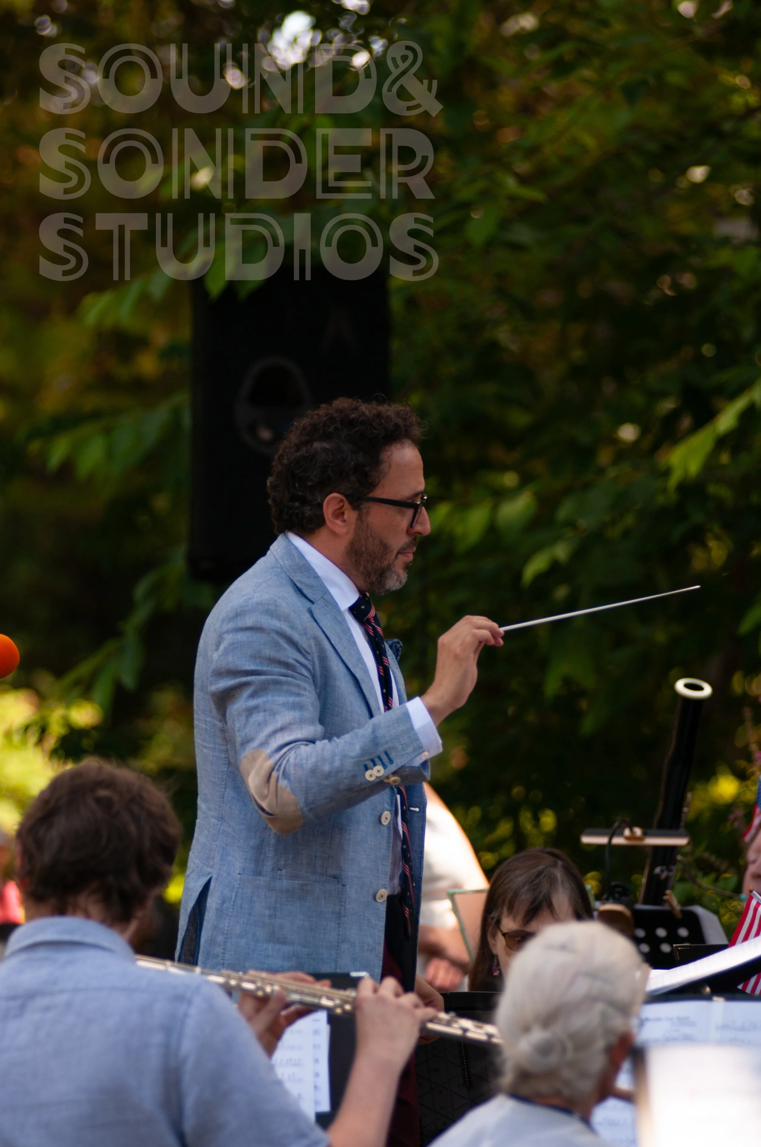 A man with curly hair and glasses wearing a light blue blazer and striped tie conducting an outdoor orchestra surrounded by musicians and seated audience members.