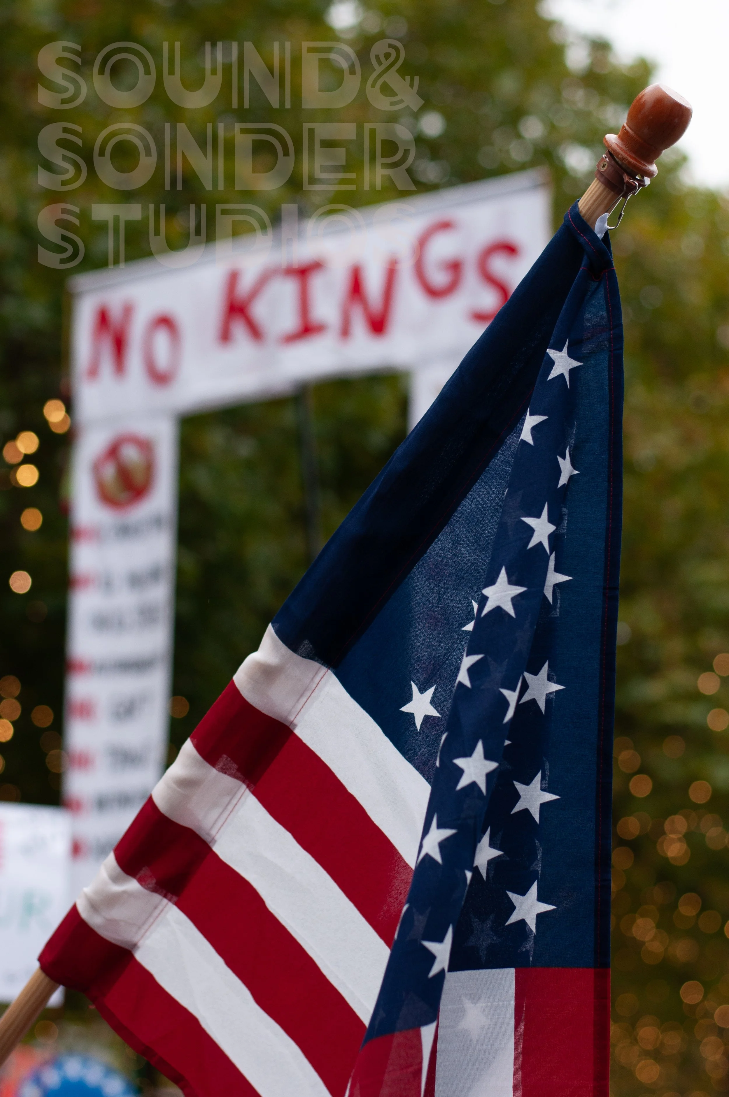 American flags with one in focus and a blurred 'No KINGS' sign in the background.
