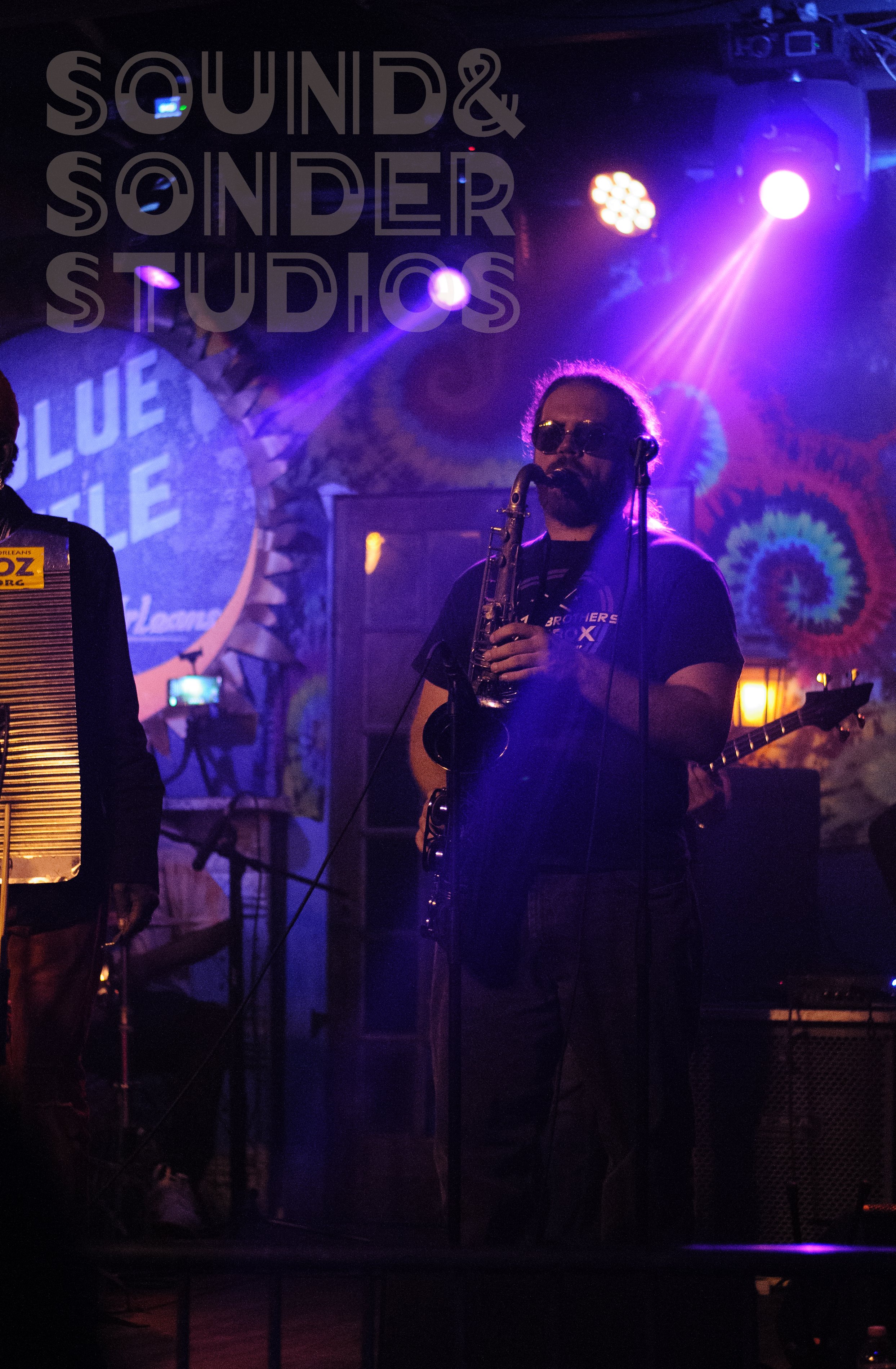 Musician wearing sunglasses playing a saxophone on stage with purple and yellow stage lights and colorful tie-dye background. Text overlay reads 'SOUND & SENDER STUDIOS' and a projection displays 'BLUE C...' on the wall.