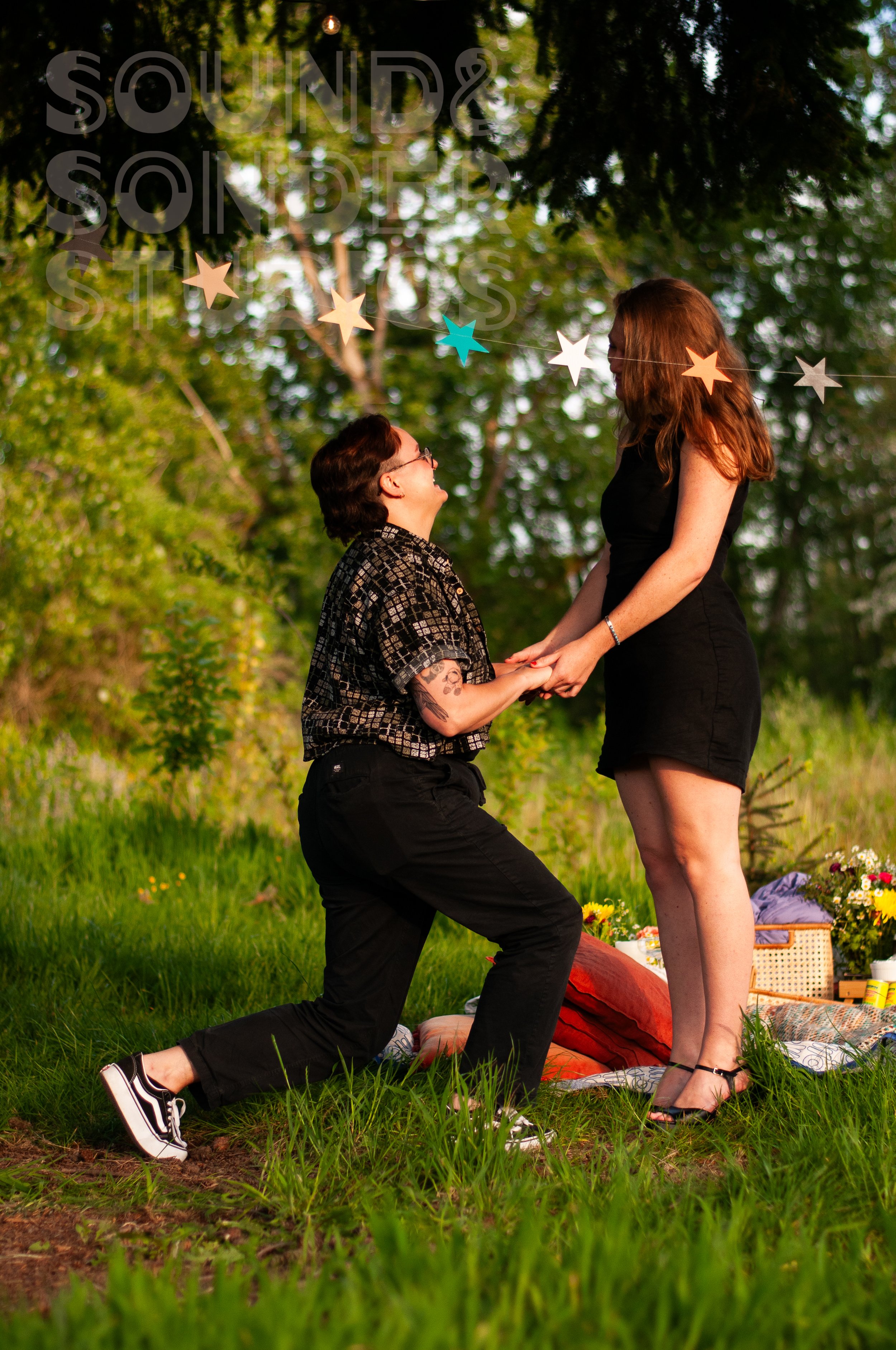 A person with short dark hair and glasses proposes marriage to a woman with long red hair outdoors. They are surrounded by trees and greenery, with a picnic setup in the background. The person on one knee holds the woman's hands, and she looks surprised.