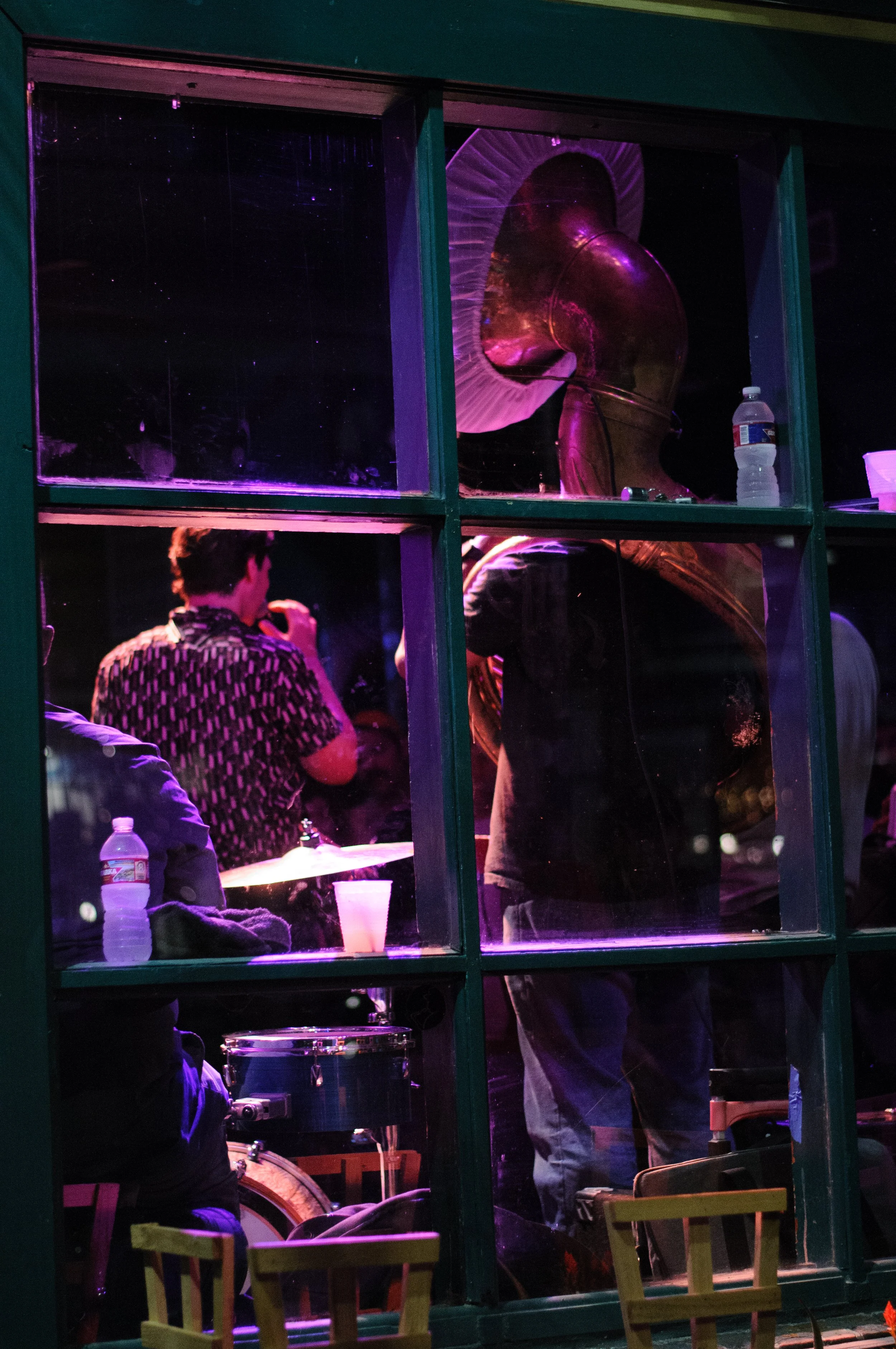 People sitting in a music venue, with a saxophone and drums visible through a window, lit with purple and pink lighting, and a water bottle placed on the windowsill.