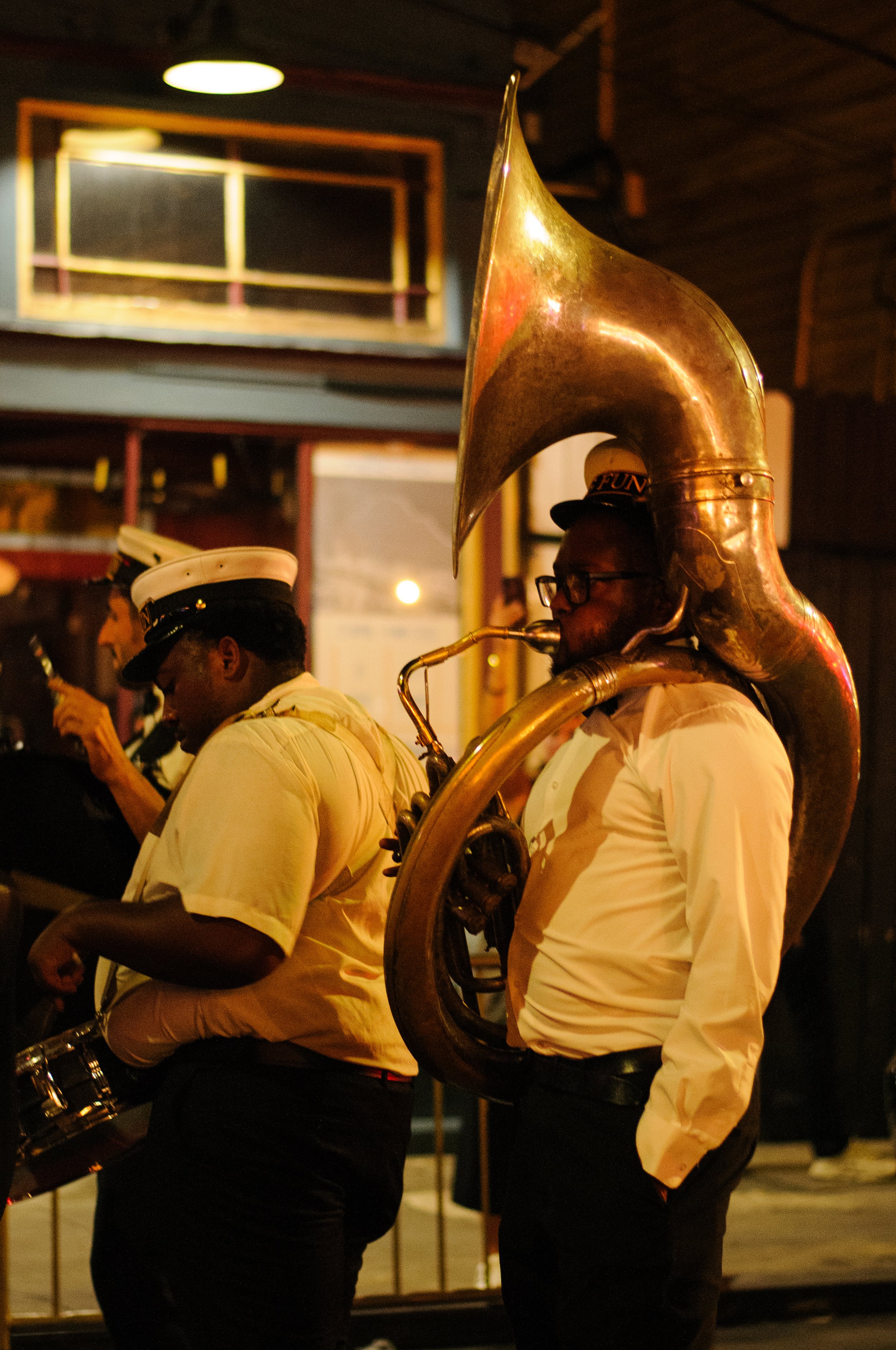 Musicians in uniform playing brass instruments inside a dimly lit room, with gold-colored instruments, including a sousaphone, and a person wearing glasses and a hat.