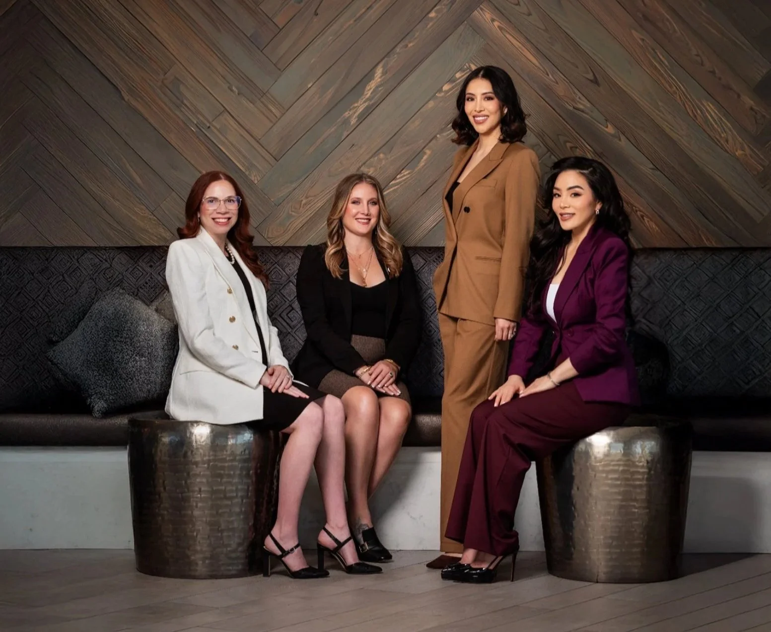 Four women dressed in professional business attire sitting and standing on a modern black leather couch with decorative pillows in front of a wooden wall.