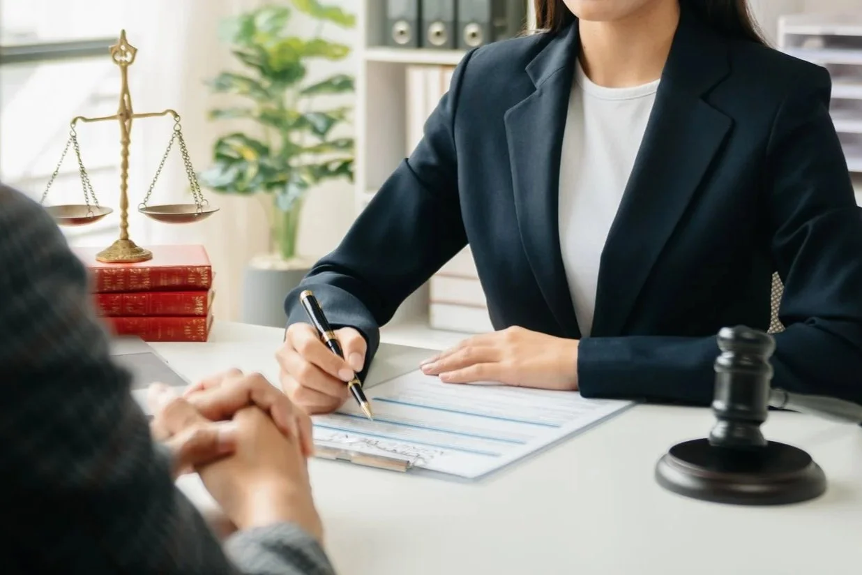 A woman in a navy blazer sitting at a desk, writing on a document during a legal consultation with a client, with a gavel and legal books on the desk and law office background.