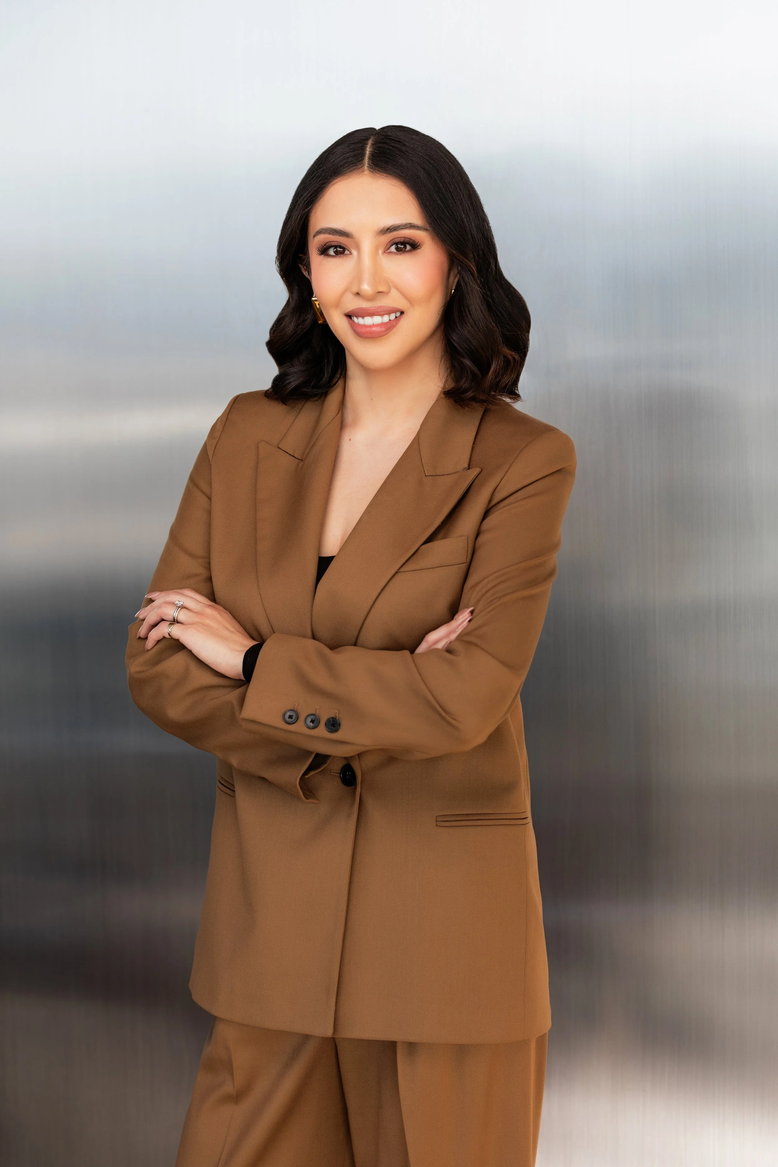 A professional woman with dark hair in loose waves, wearing a brown blazer and matching pants, standing with folded arms against a metallic background.