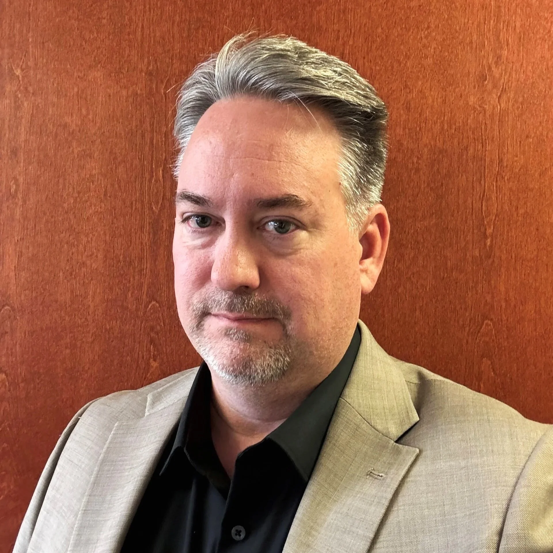 A man with short gray hair, wearing a beige blazer over a black shirt, standing against a reddish-brown background.
