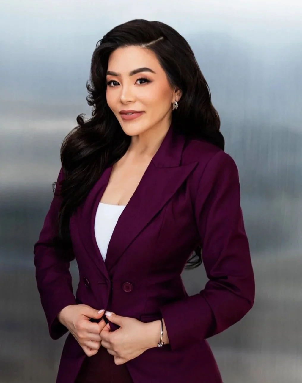 A woman with long dark wavy hair wearing a burgundy blazer and a white top, standing against a blurred metallic background.