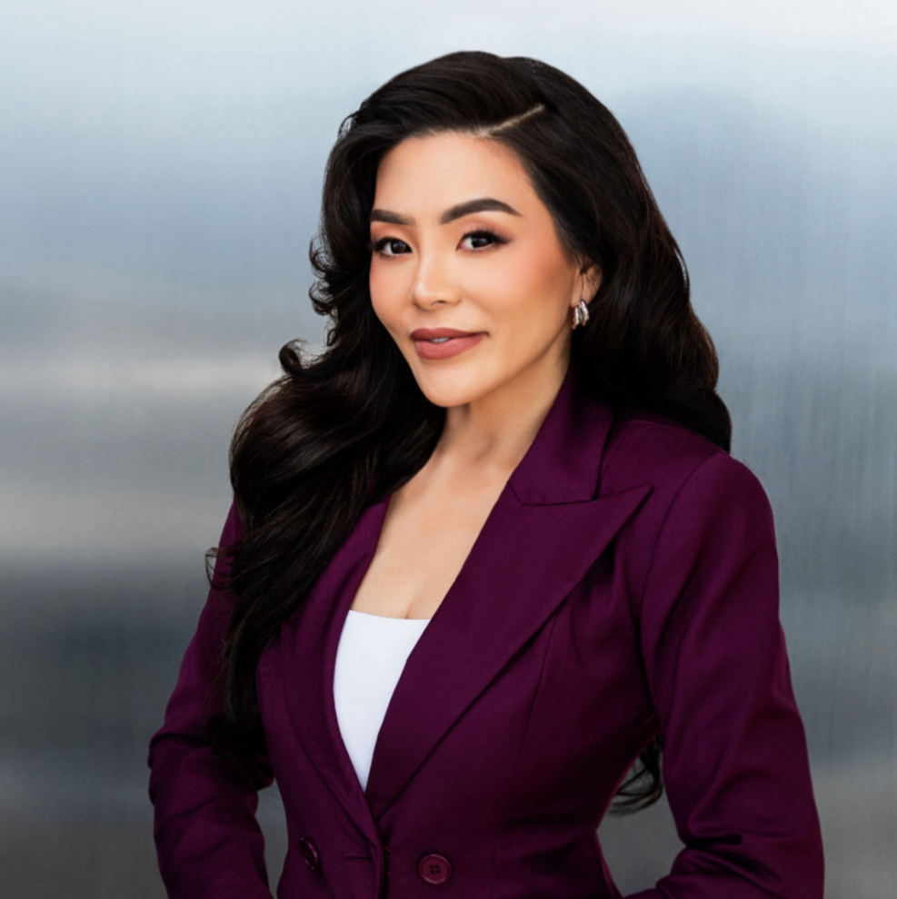 A woman with long, dark wavy hair wearing a purple blazer and earrings, posing in front of a gray background.