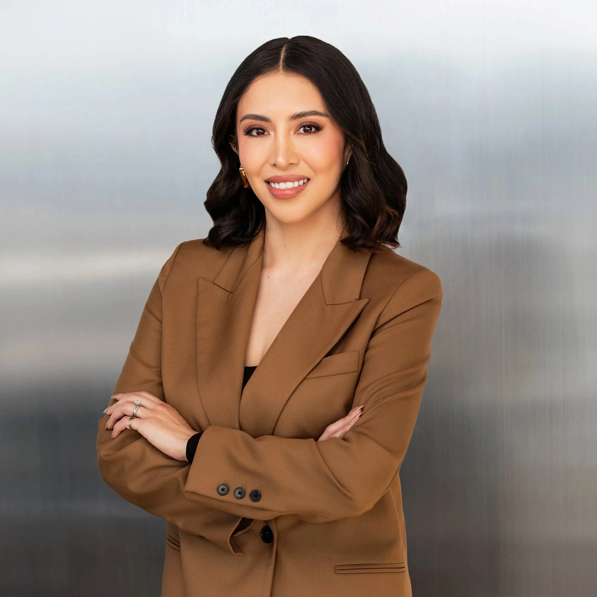 A woman with dark wavy hair, wearing a brown blazer and gold earrings, is smiling and crossing her arms in front of a neutral gradient background.