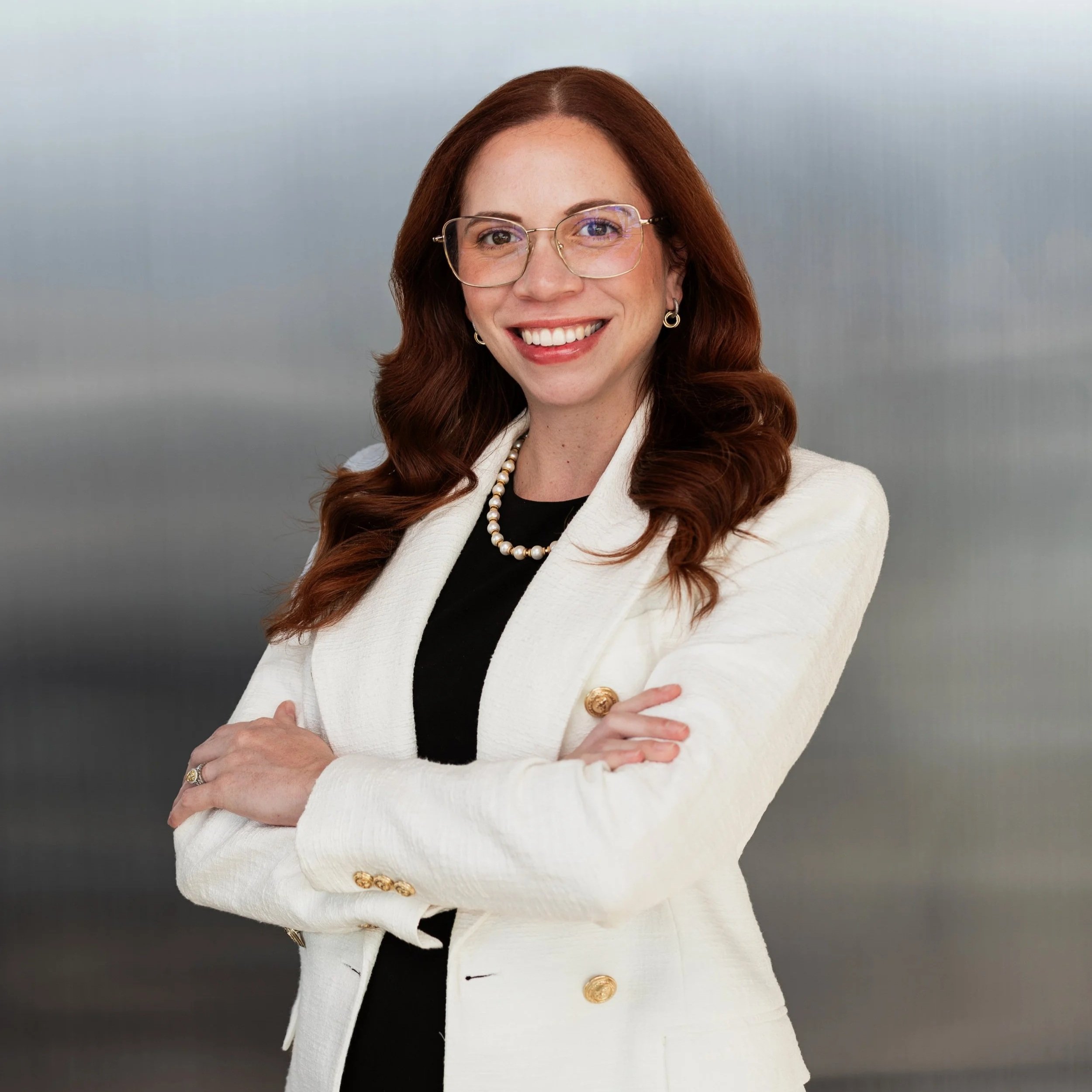 A woman with long red hair, glasses, earrings, and a pearl necklace, smiling and wearing a white blazer over a black top, with crossed arms, against a plain light-colored background.