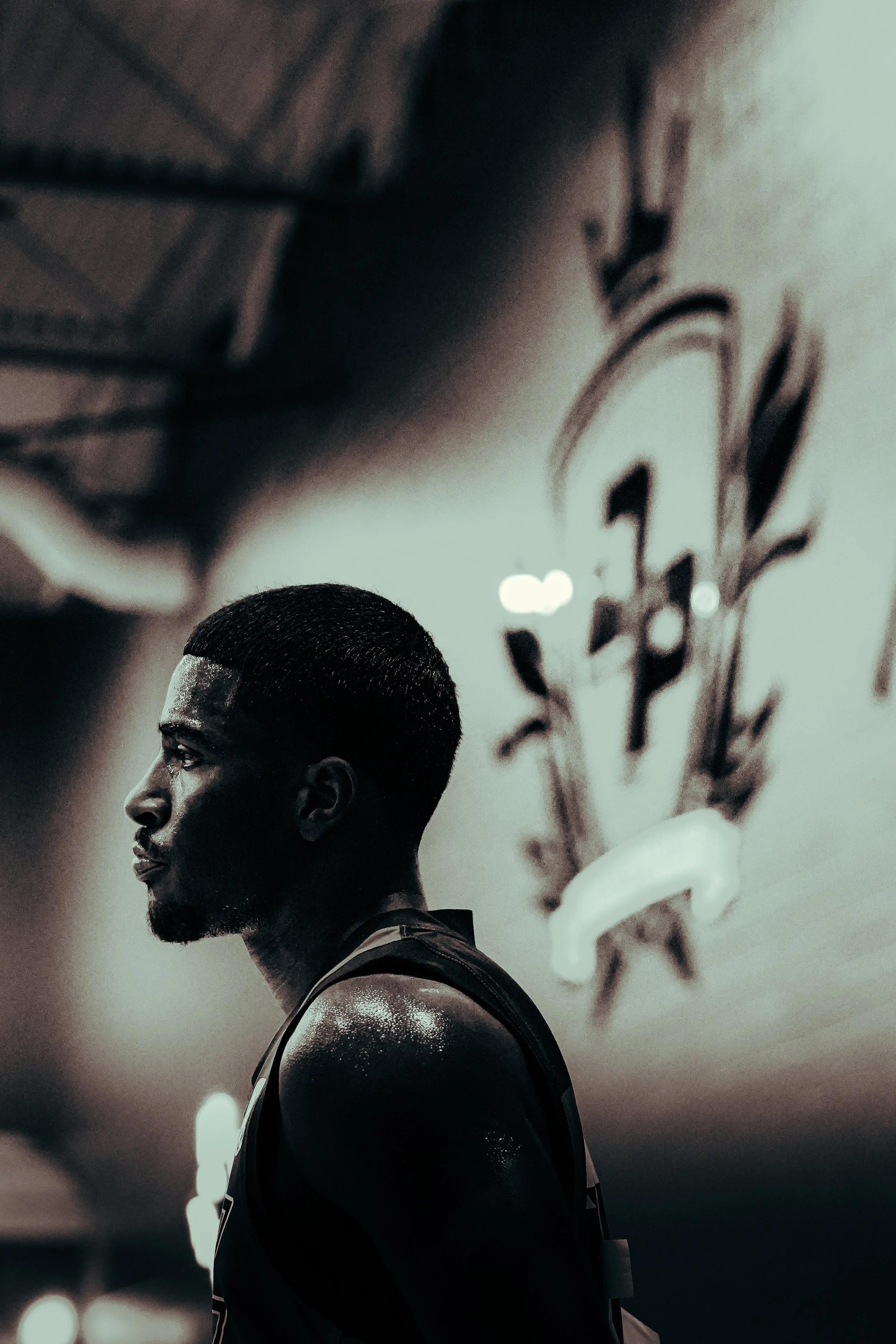 A male athlete with a short haircut and beard, sweating and wearing a sleeveless sports jersey, standing indoors with a blurred background that has a large graffiti-style artwork on the wall.