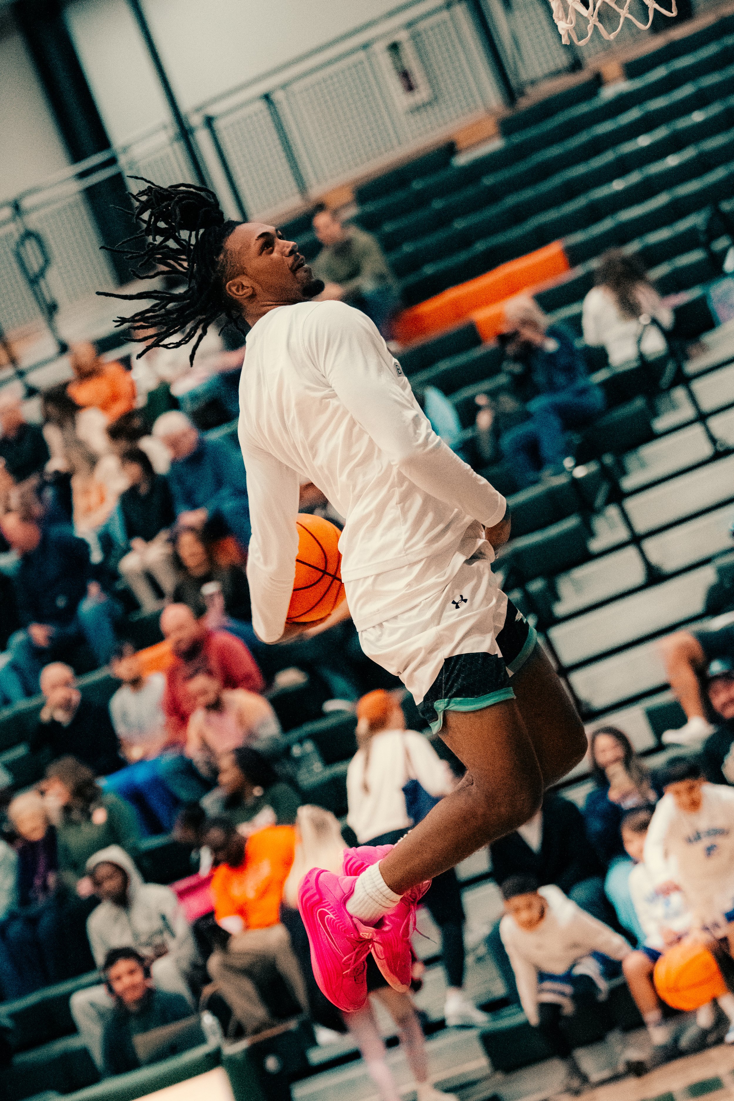 A female basketball player with dreadlocks wearing a white shirt and black shorts, jumping to make a shot with a basketball in a gymnasium filled with spectators.